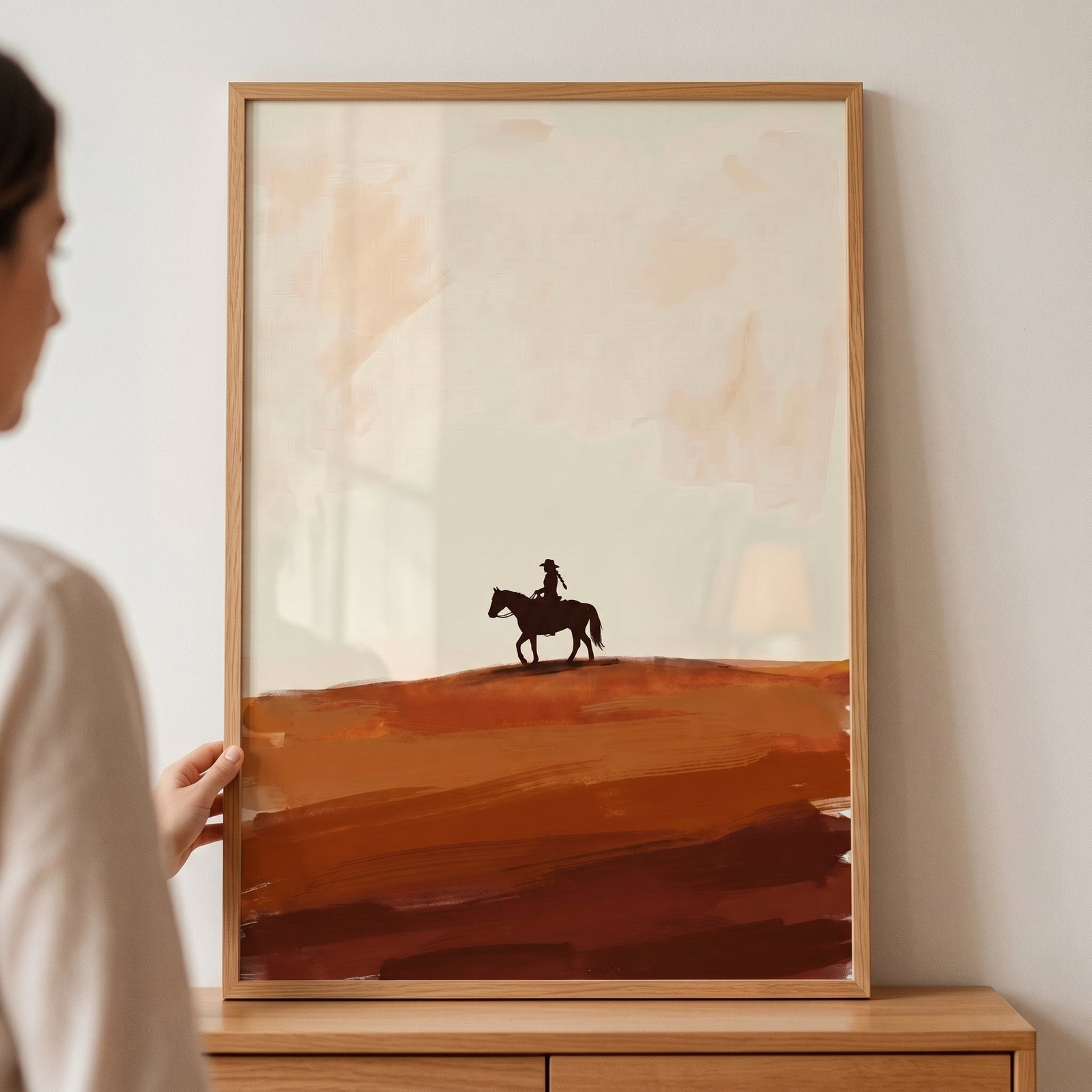 Minimalist warm horizon cowboy wall art in a light wood frame shows a horse and rider silhouette on layered earth tones.