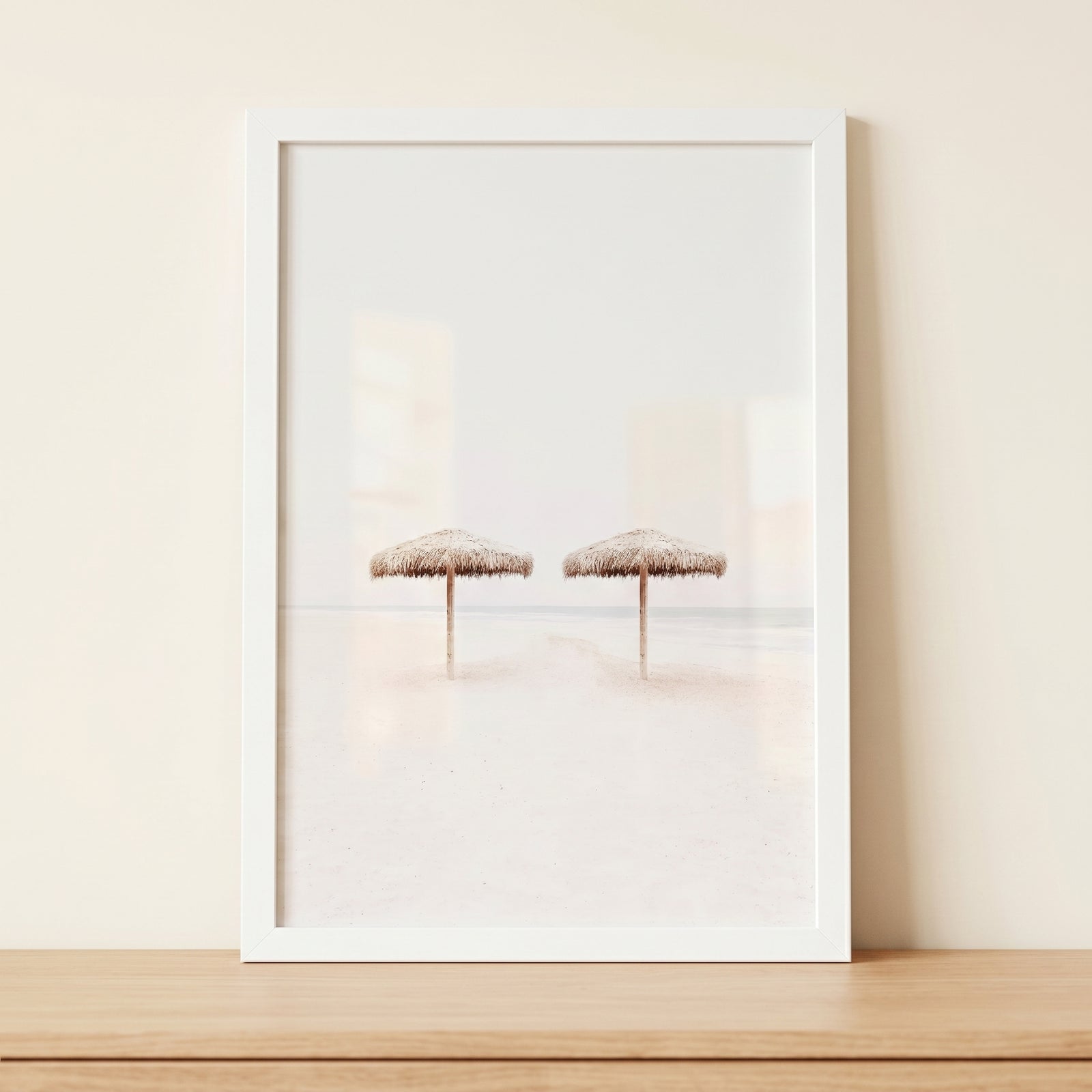 Minimalist white beach umbrella wall art shows two straw umbrellas on a bright beach, framed and resting on a wood shelf.