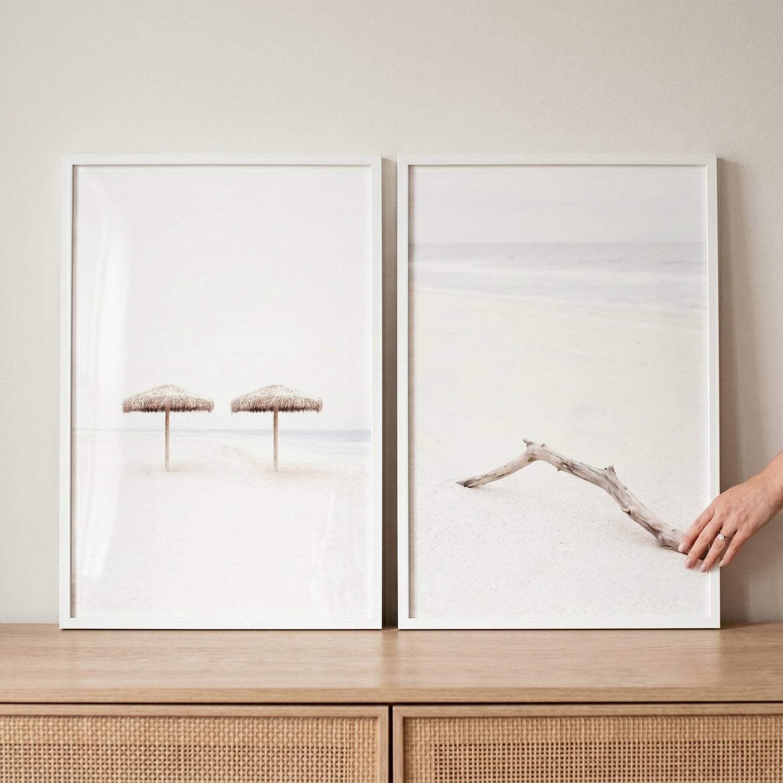 Minimalist white sand beach wall art set of 2 featuring straw umbrellas and driftwood, with a hand touching one frame.