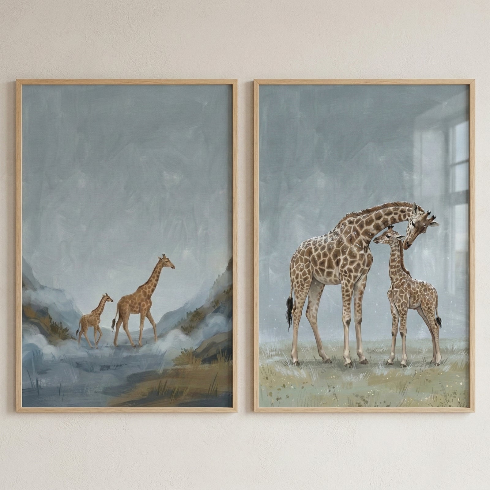 Two framed pieces of art featuring giraffes are hung side-by-side on a light wall. Each piece is set in a thin, light-wood frame. The left image shows an adult and a young giraffe walking through a misty landscape with hills and a blue shoreline. The right image depicts an adult giraffe tenderly nuzzling a smaller one on a grassy plain under a soft, blue-grey sky. The style is gentle and illustrative, with a muted color palette of blues, greens, and browns.