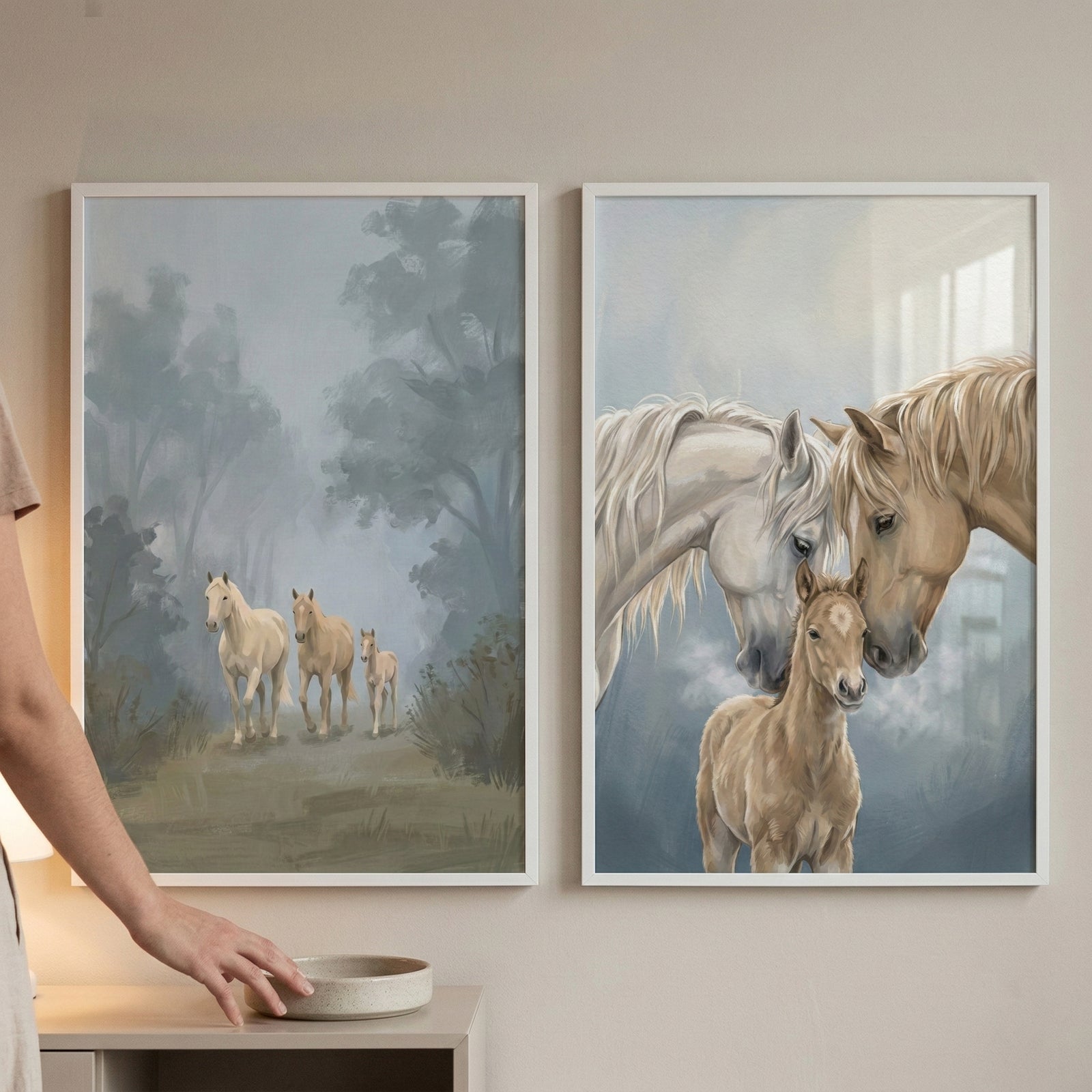 Two framed horse prints hang on a beige wall above a light surface, with part of a person visible at the left for scale. One print shows three pale horses walking through a misty wooded path, while the other shows two adult horses nuzzling a foal against a soft blue gray background.