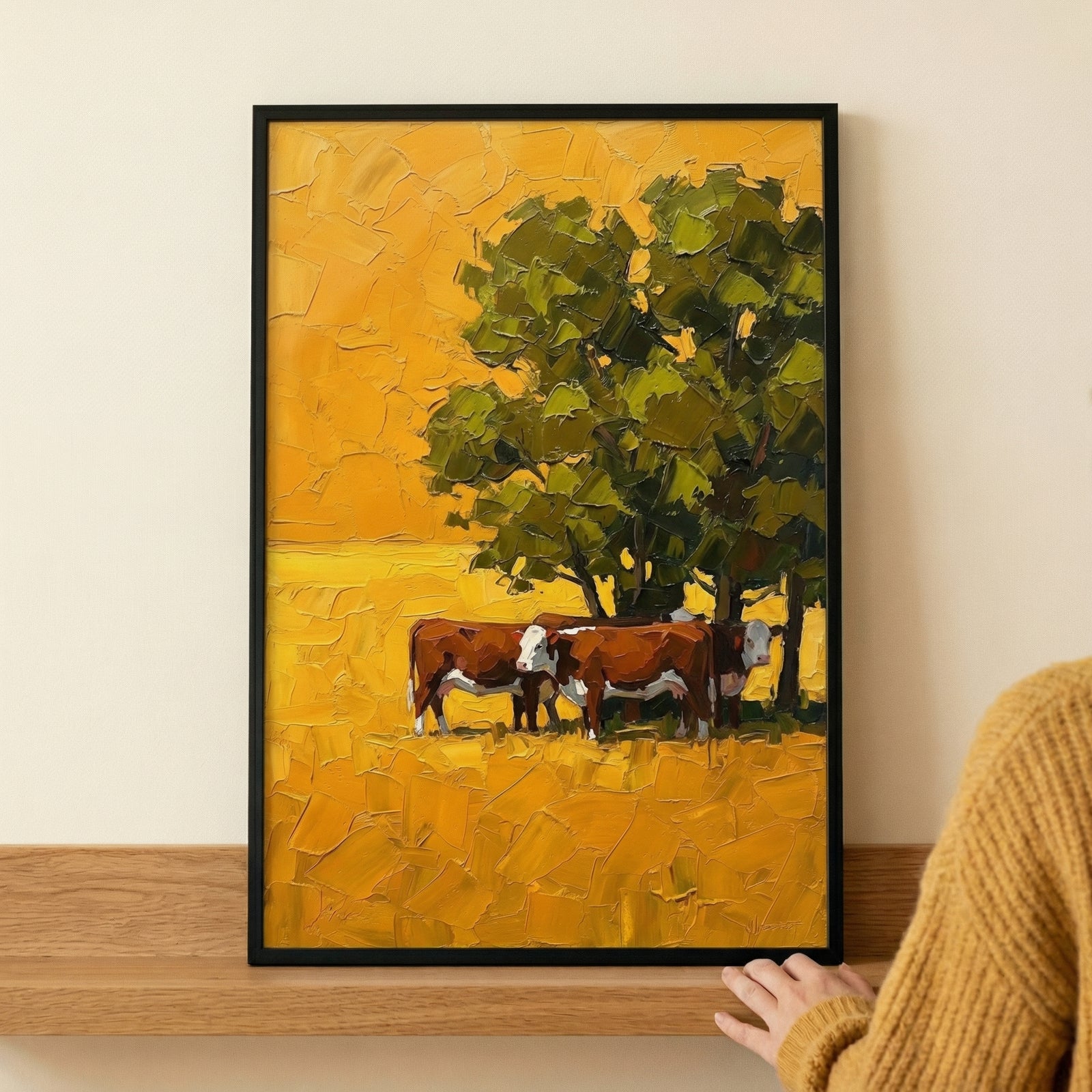 A vertical framed artwork sits atop a light-toned wooden shelf against a plain wall. The piece displays a vibrant, impressionistic rural scene rendered with thick, textured strokes in shades of golden yellow, amber, and deep green. In the center, three brown and white cows stand together beneath the canopy of a large, leafy tree. The surrounding field and sky are composed of bold, heavy layers that create a rich surface quality. 