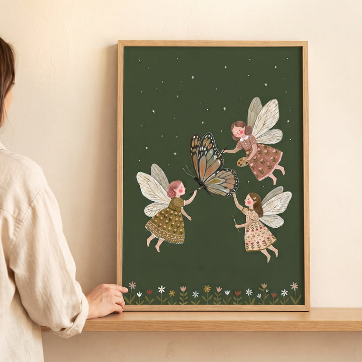 Monarch butterfly artist fairies wall art shows three winged figures decorating a butterfly against a starry green sky.