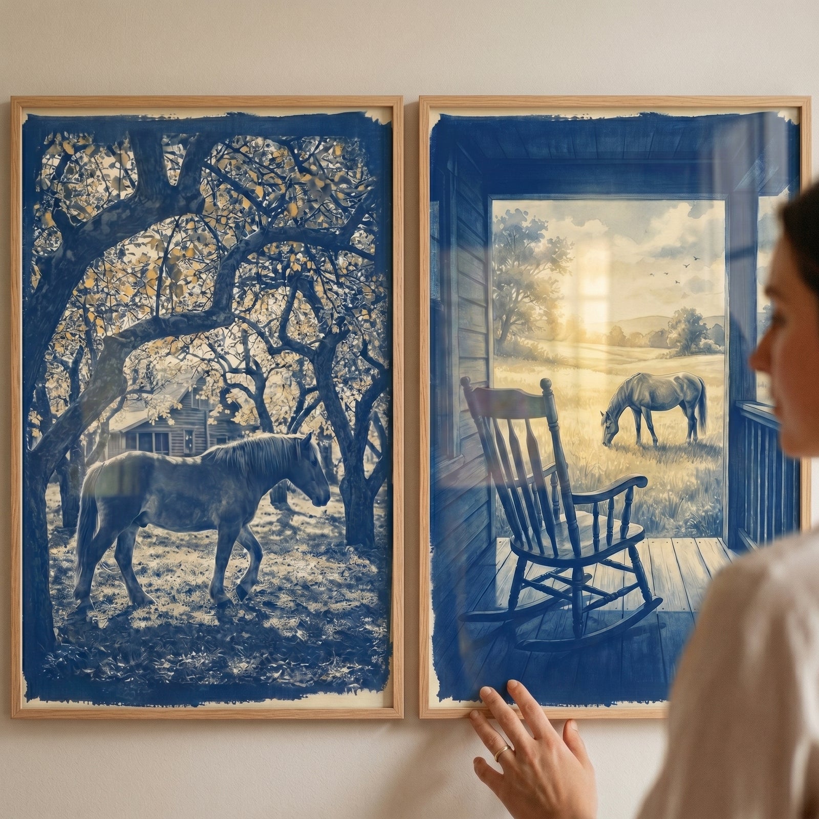 A person adjusts a monochrome blue horse wall art set of 2 featuring rural landscapes on a neutral wall.