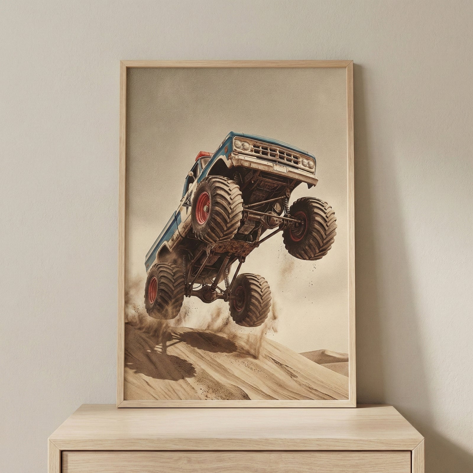A framed vertical print featuring a blue vintage monster truck with oversized, knobby tires leaping off a sand dune. The truck is angled sharply upward mid-jump, kicking up dust against a vast desert background and a hazy, neutral sky. The artwork is encased in a light wood frame resting on a wooden cabinet against a plain wall.