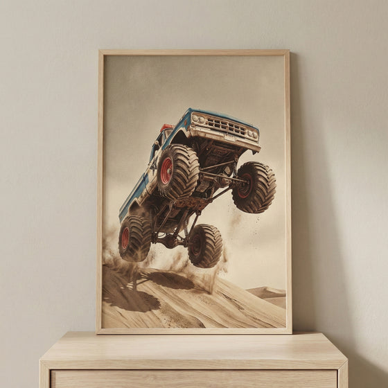 A framed vertical print featuring a blue vintage monster truck with oversized, knobby tires leaping off a sand dune. The truck is angled sharply upward mid-jump, kicking up dust against a vast desert background and a hazy, neutral sky. The artwork is encased in a light wood frame resting on a wooden cabinet against a plain wall.