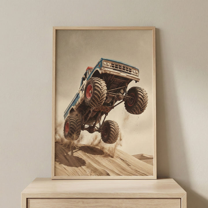 A framed vertical print featuring a blue vintage monster truck with oversized, knobby tires leaping off a sand dune. The truck is angled sharply upward mid-jump, kicking up dust against a vast desert background and a hazy, neutral sky. The artwork is encased in a light wood frame resting on a wooden cabinet against a plain wall.