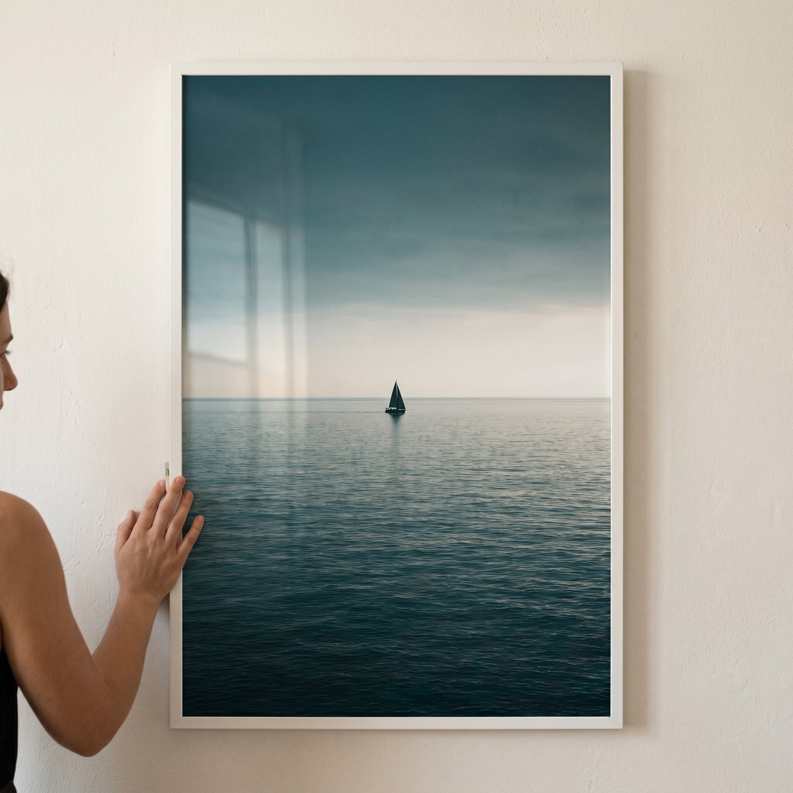 Hand touching a moody teal sea sailboat wall art showing a lone boat on a vast, dark ocean under a cloudy sky.