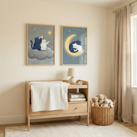 A serene, neutral-toned nursery features two framed illustrations above a light wood changing table. In one illustration, a black cat and a white cat sit on a fluffy cloud, looking through a telescope at a single bright star. The second shows the same two cats curled up and sleeping on a glowing yellow crescent moon. A small white lamp and folded blankets rest on the changing table. A wicker basket overflowing with plush rabbit toys sits on a soft white rug beside the furniture.