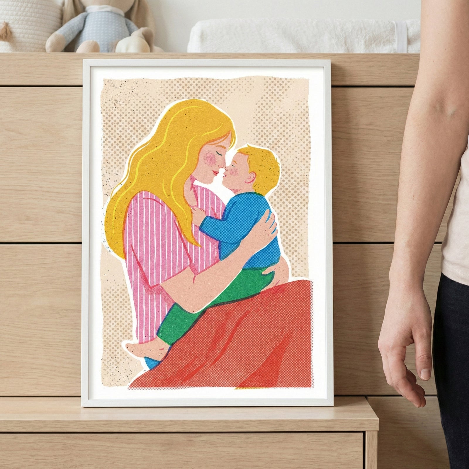 A vertical framed illustration sits on a light wooden surface, depicting a tender moment between a woman and a young child. The woman, shown with long, flowing yellow hair, wears a pink and white striped top and holds the child closely. The child has short yellow hair and is dressed in a blue long-sleeved shirt and green pants. They are both shown in profile, their faces nearly touching in an affectionate embrace.
