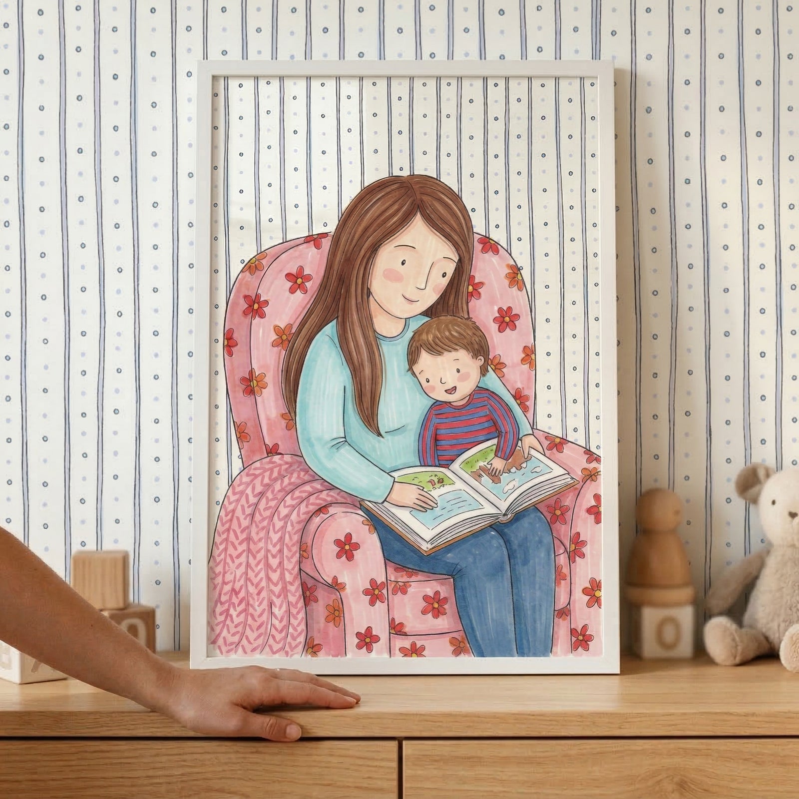 This framed piece of art features a heartwarming illustration of a mother and her young child sharing a quiet moment together. The woman, with long brown hair and a gentle expression, sits in a pink armchair decorated with a red floral pattern. She wears a light blue sweater and dark blue trousers. On her lap, a small child in a red and blue striped shirt is engrossed in an open picture book they are reading together. A pink blanket is draped over one arm of the chair. 
