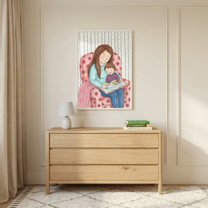 A cozy interior scene featuring a vertical framed illustration centered on a neutral-toned wall with elegant molding. The artwork depicts a mother with long brown hair sitting in a pink floral armchair, reading a book to a young child on her lap. The illustration is done in a soft, whimsical, watercolor-like style. Below the framed print, a light-colored wood dresser with three drawers holds a small decorative lamp with a patterned shade on one side and a neat stack of books on the other. 