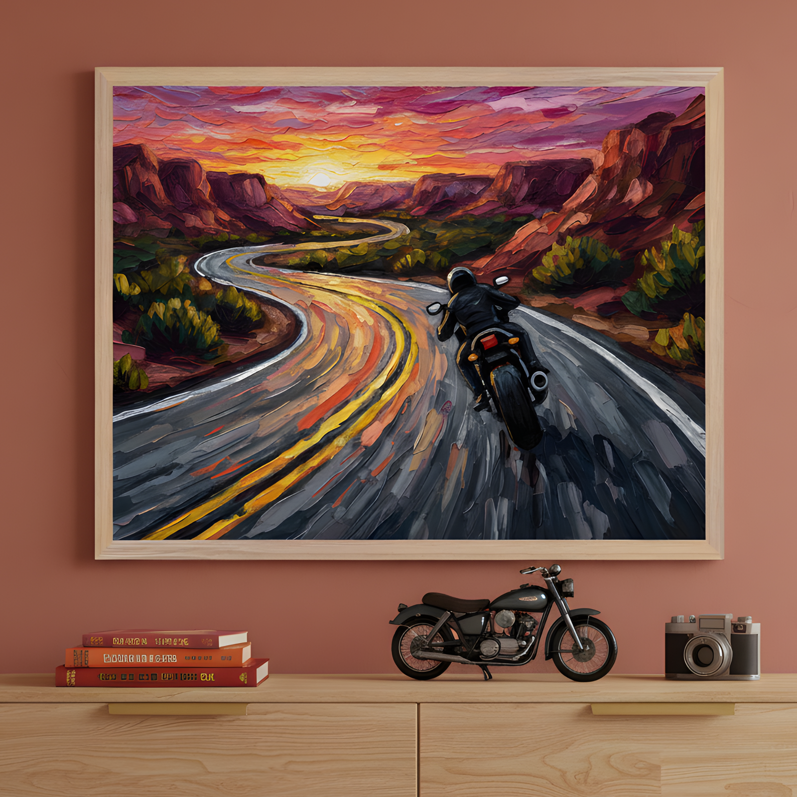 Framed artwork of a motorcyclist riding down a winding road at sunset hangs on a terracotta-colored wall. Below it, a wooden shelf displays a miniature motorcycle, a vintage camera, and stacked books with red and orange spines, echoing the warm tones of the artwork.