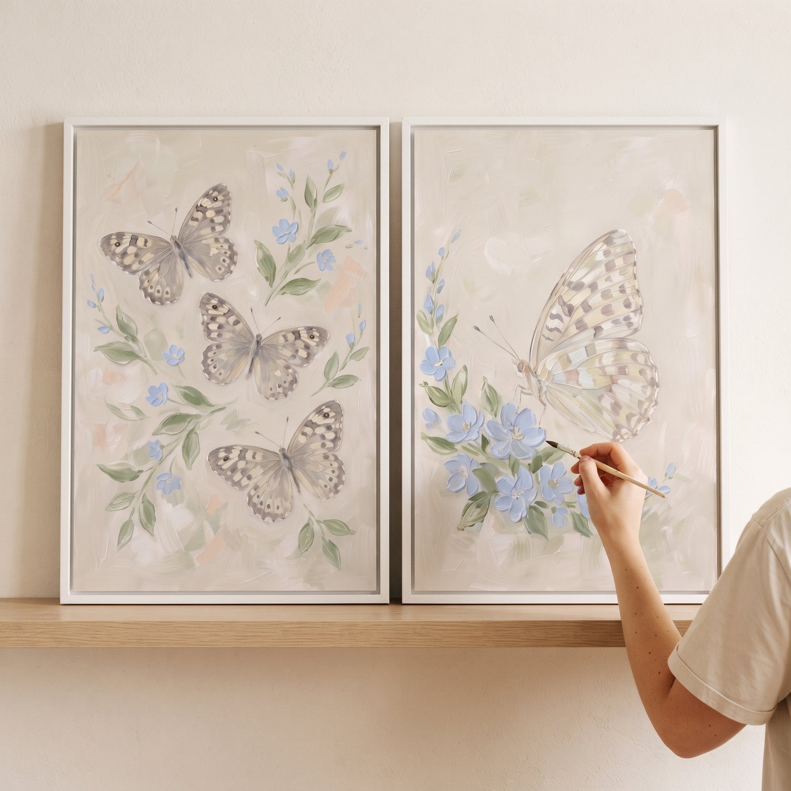 Mottled wing botanical butterfly wall art set of 2 with blue flowers and butterflies on a shelf with a hand holding a brush.