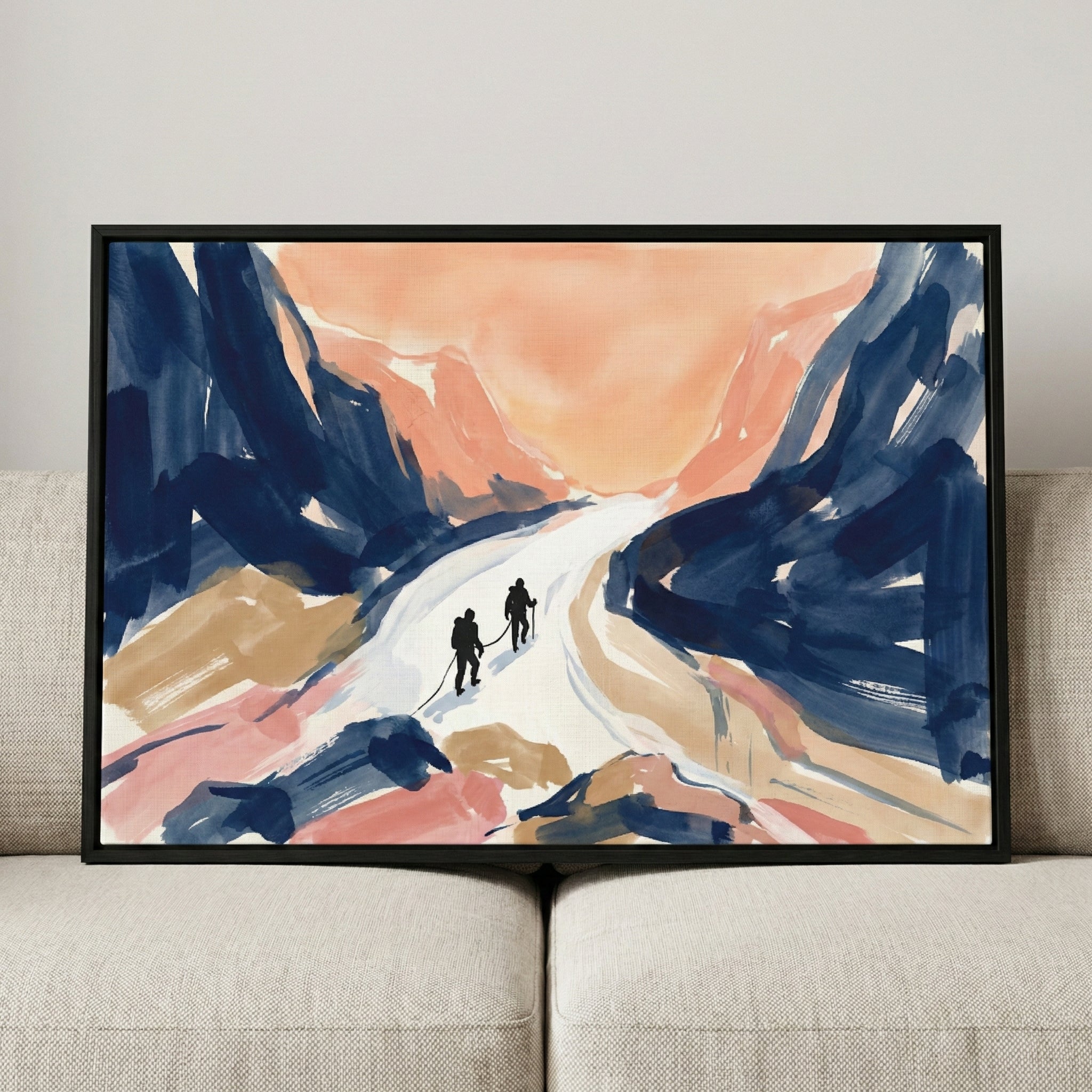An expressive artwork in a black frame depicts two roped-together silhouetted figures trekking across a bright white snowy path. The surrounding abstract mountains are rendered in broad strokes of navy blue, soft peach, and tan. The piece is resting on a light-colored fabric sofa.