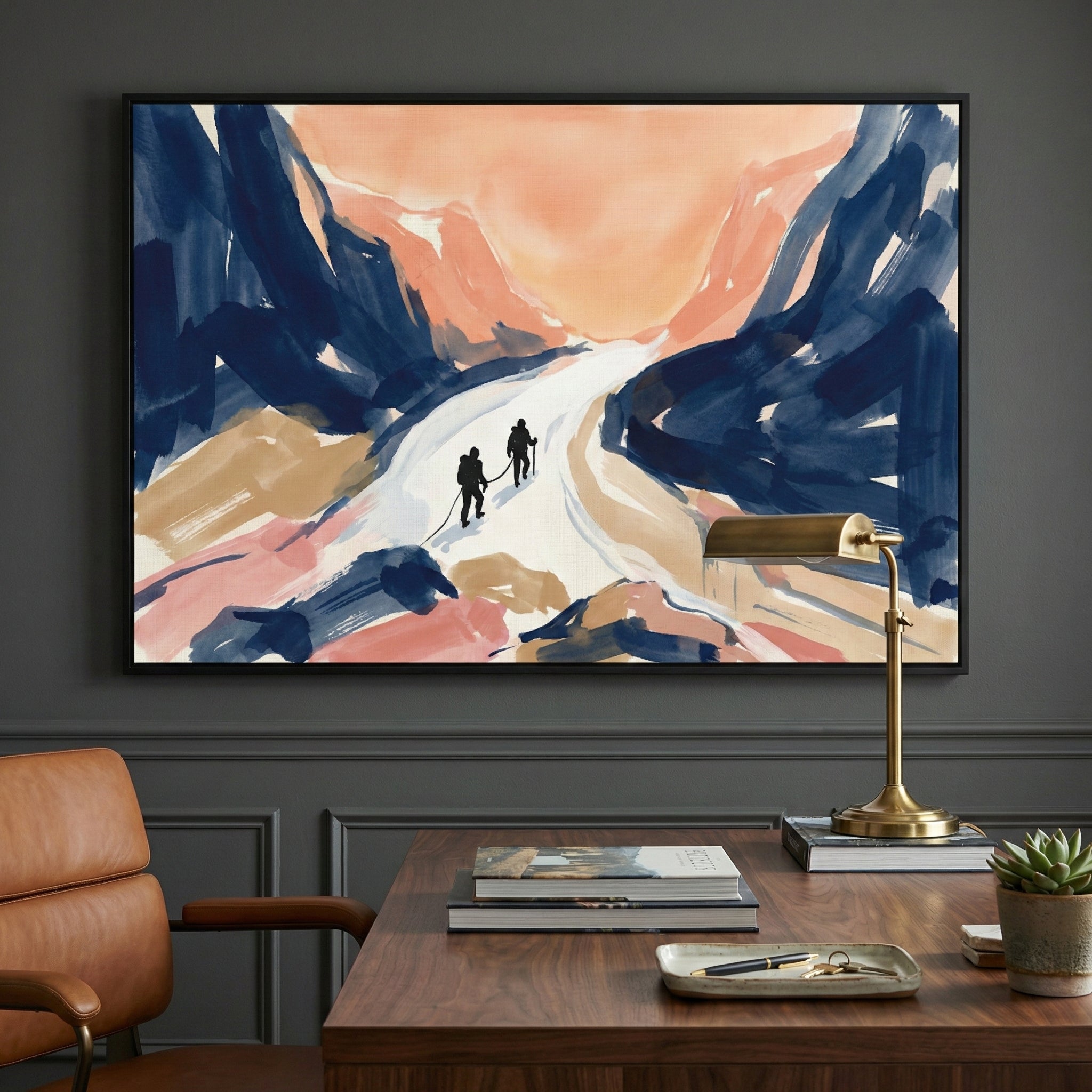 A framed abstract illustration hangs on a dark charcoal wall above a wooden desk. The artwork features two small black silhouettes of figures trekking along a white path through a mountain landscape, rendered with broad, expressive brushstrokes in navy blue, peach, tan, and soft pink. On the desk below, there is a stack of books, a brass desk lamp, a small tray with a pen, and a potted succulent. A portion of a brown leather chair is visible on the left side of the frame.