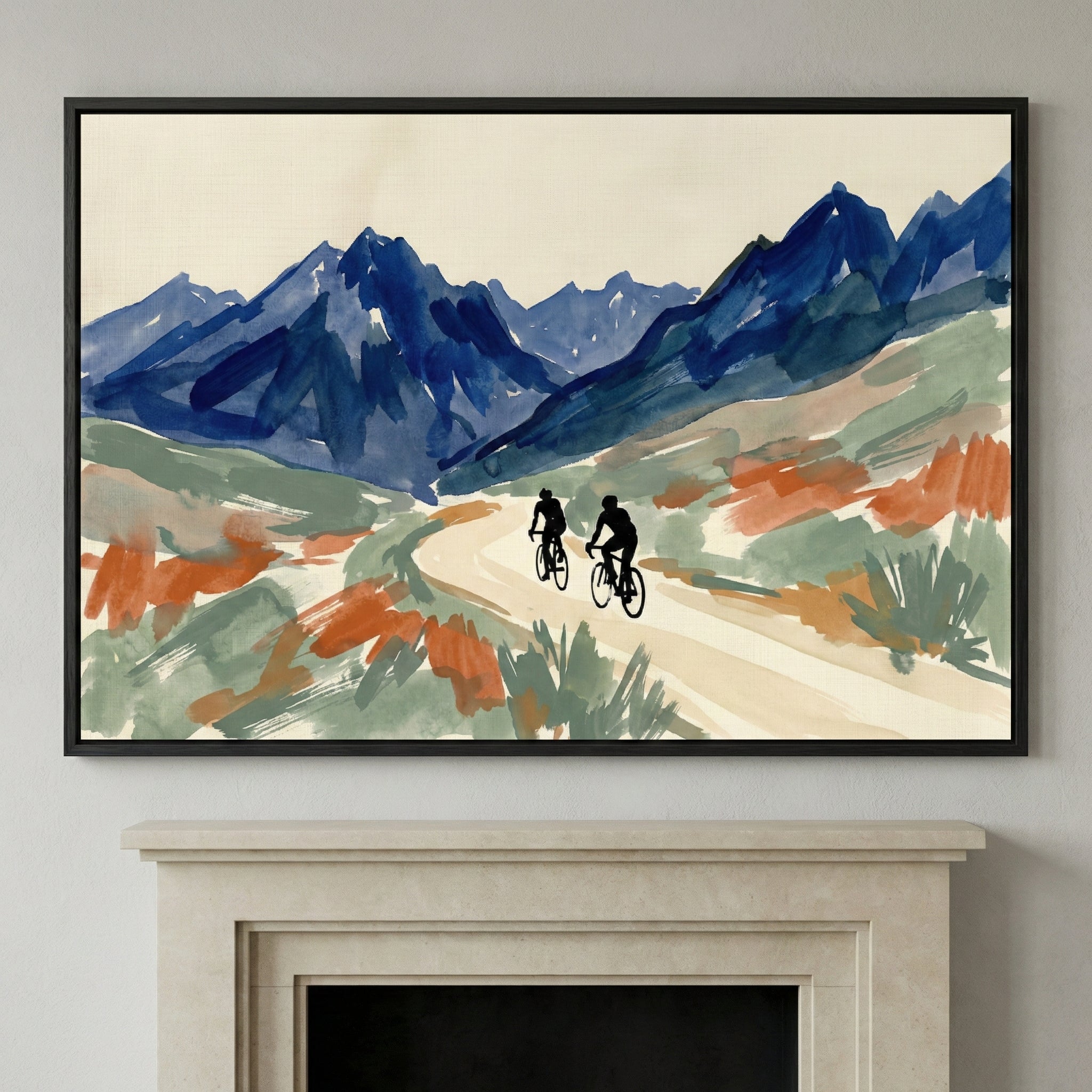 An expressive art piece in a slim black frame hangs on a neutral-toned wall above a light stone fireplace mantel. The artwork depicts two silhouetted figures on bicycles traveling along a winding, light-colored path. In the background, a range of deep blue, jagged mountains stands against a pale cream sky. The surrounding landscape is rendered with loose, gestural brushstrokes in shades of sage green and terracotta orange, suggesting desert vegetation.