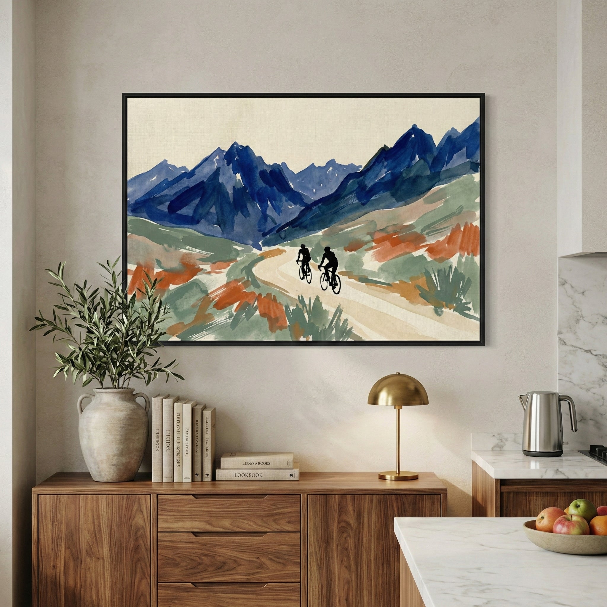 A stylish interior room with a large framed landscape illustration hanging on a beige wall. The scene in the frame shows two cyclists riding on a light-colored winding path through a valley, with tall blue mountains in the distance and patches of green and terracotta-colored brushstrokes in the foreground. Beneath the frame, a mid-century style wooden cabinet is decorated with a leafy green plant in a light-colored vase, a neat row of books, and a gold table lamp. 