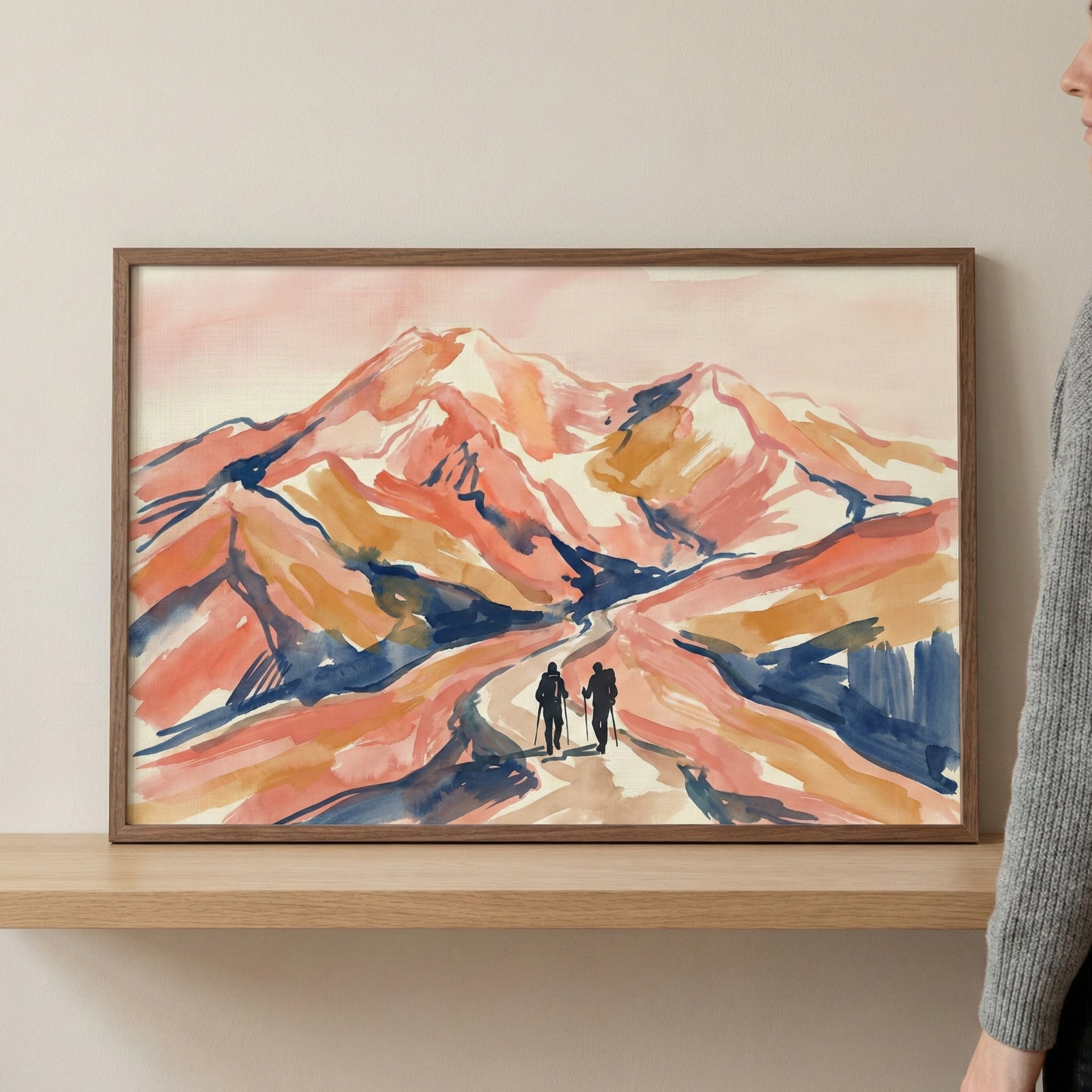 A framed piece of artwork depicting a mountain landscape in a loose, expressive style is shown resting on a light wood shelf. The scene features mountain peaks rendered in warm shades of coral, orange, and peach, contrasted with deep navy blue shadows and white highlights. Two small silhouetted figures with backpacks and trekking poles are seen walking on a path that winds into the mountains. The dark wood frame is set against a plain, neutral wall.