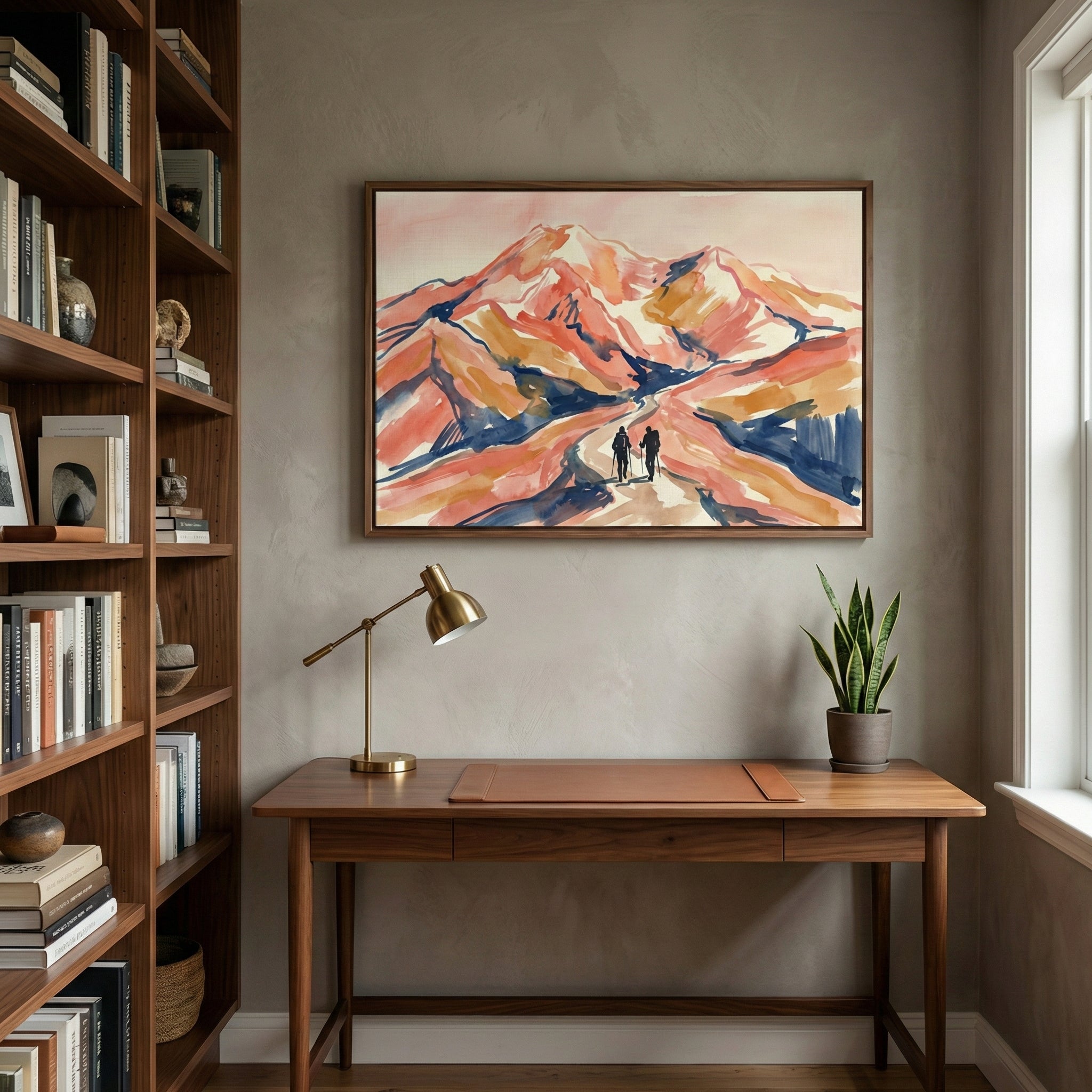 A bright home office features a wooden desk centered against a textured gray wall. Above the desk hangs a large framed horizontal artwork depicting an abstract mountain landscape in shades of peach, orange, and deep blue, with two small figures walking along a trail. On the desk sit a slim brass lamp, a large leather desk pad, and a small potted green plant. To the left is a tall wooden bookshelf filled with books and decorative objects. 