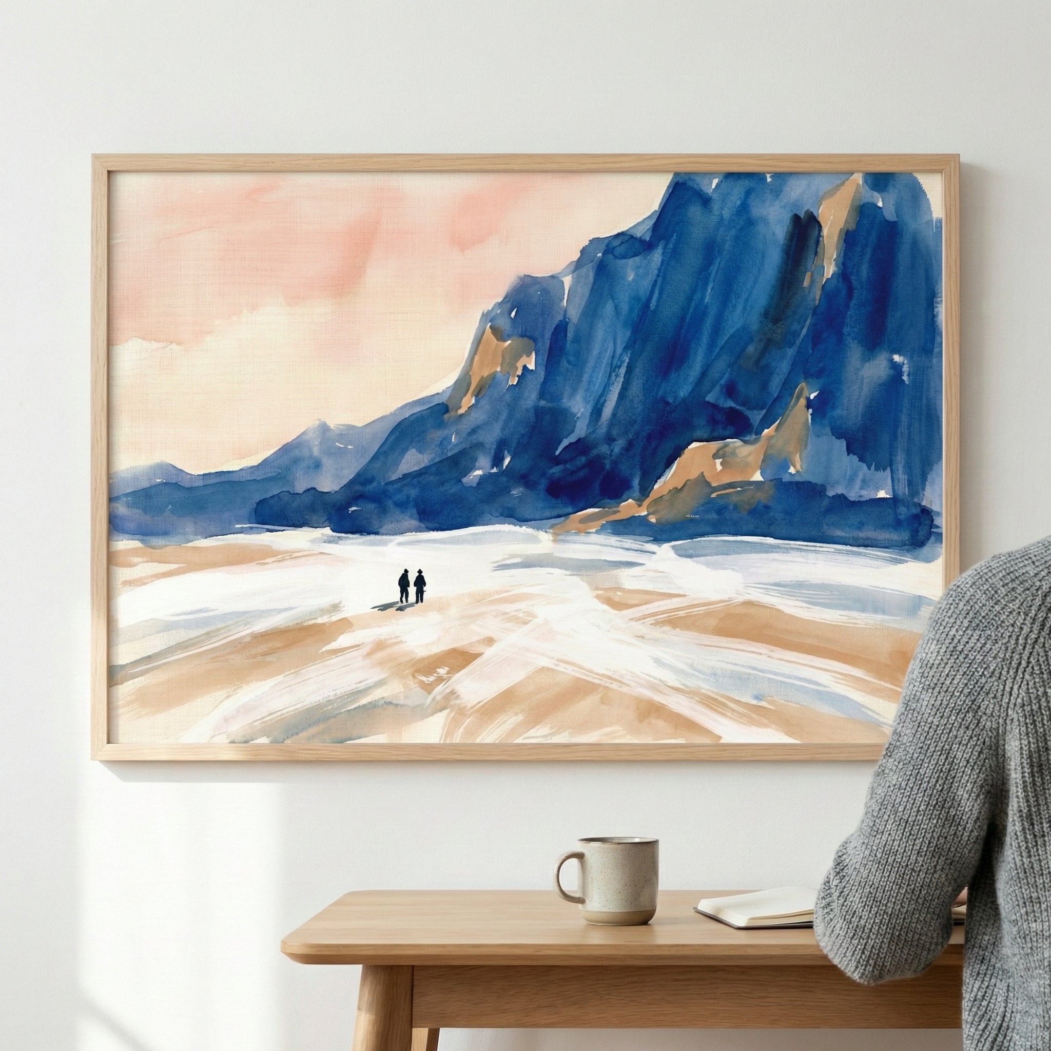 A framed piece of landscape art hangs on a plain white wall above a wooden desk. The artwork depicts a large, dark blue mountain or cliff under a soft peach-colored sky. In the foreground, two small silhouettes of people walk across a broad, textured landscape of white and tan. Below the frame, a ceramic mug and a notebook sit on a light wood desk. The shoulder and arm of a person wearing a grey sweater are visible in the foreground on the right.