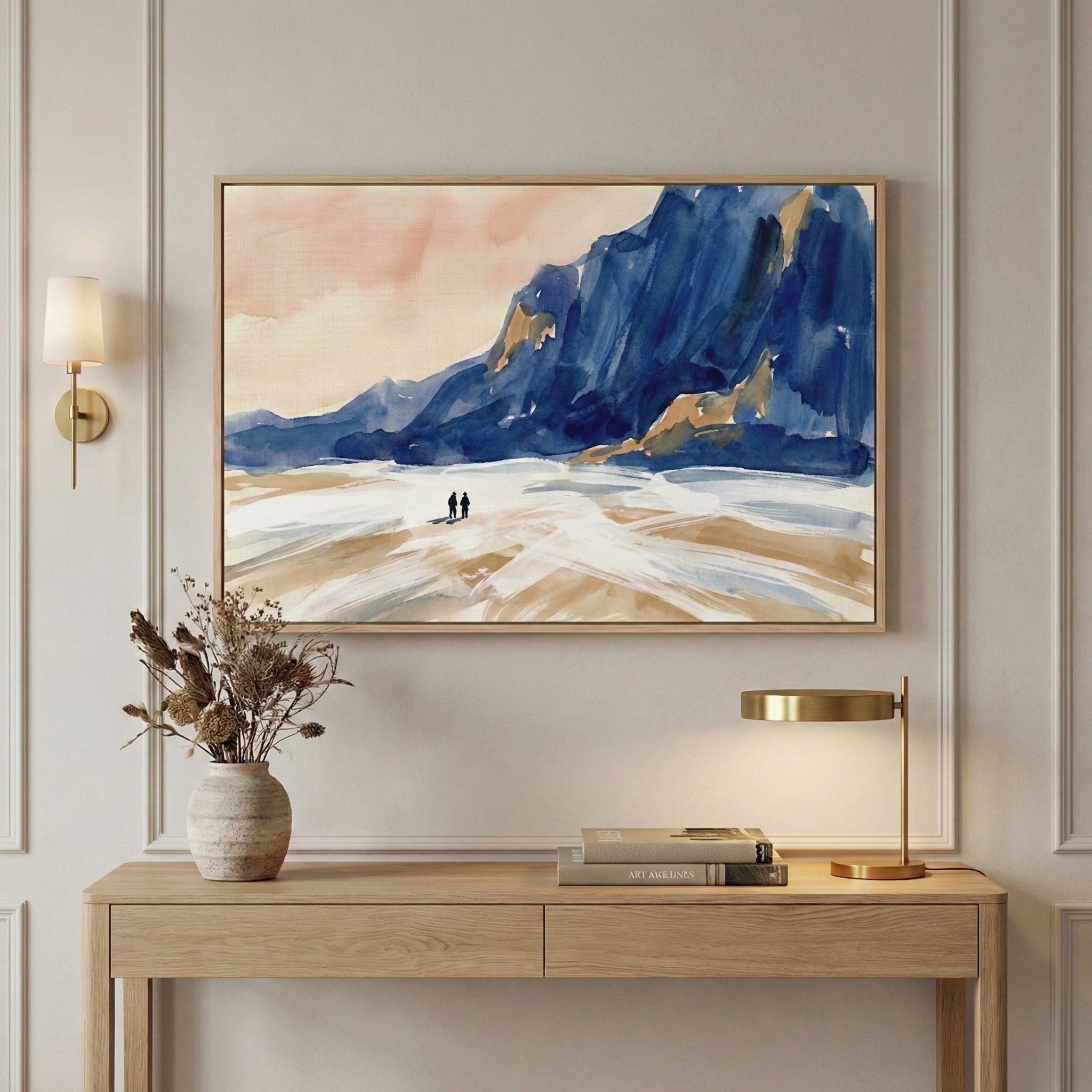 A minimalist interior scene featuring a large framed piece of landscape artwork centered on a light-colored wall with decorative molding. The visual piece depicts an abstract blue mountain range on the right against a soft peach and white sky, with two small dark silhouettes standing in a white and sandy-colored foreground. Below the framed artwork is a light wood console table. On the left side of the table sits a textured beige ceramic vase filled with dried botanicals.