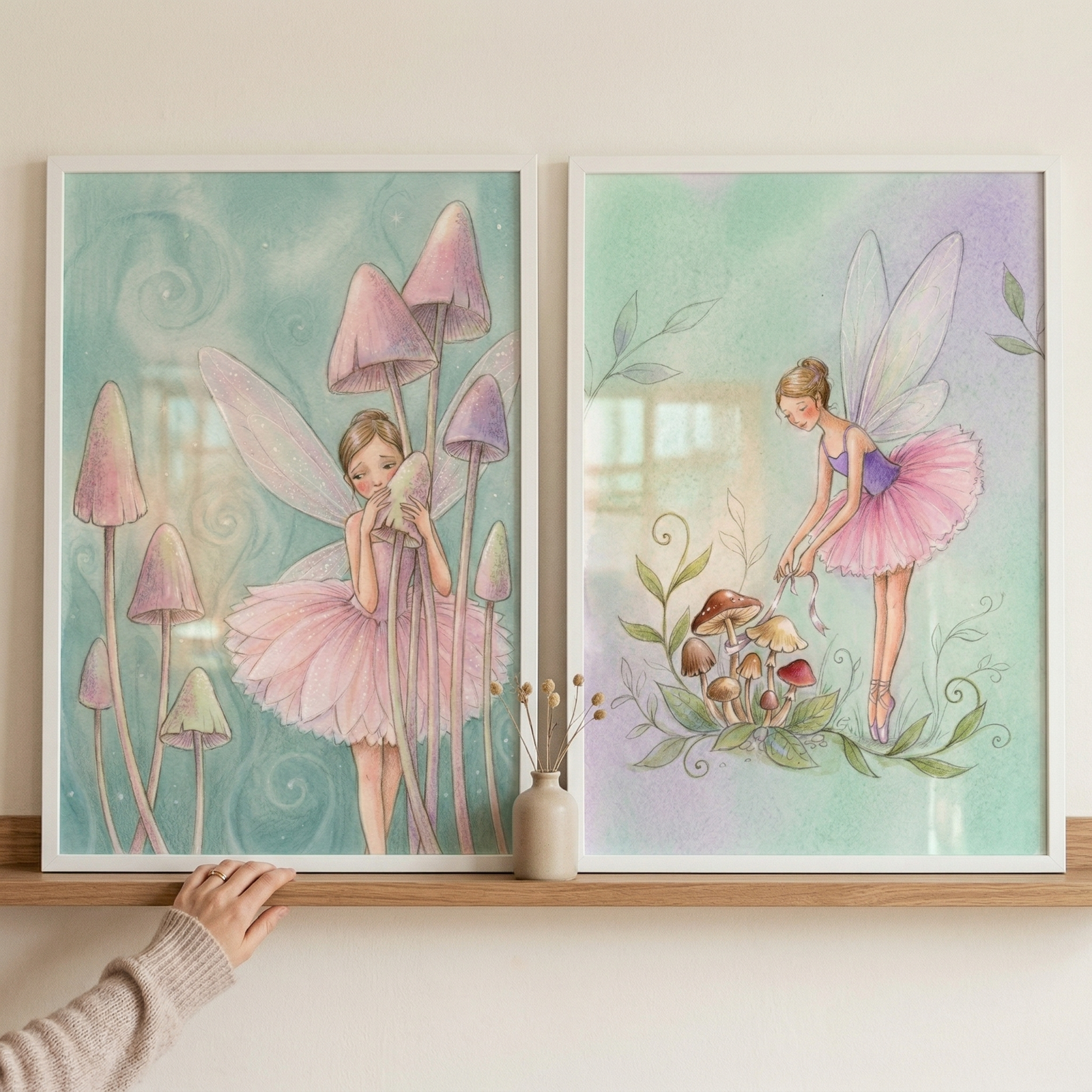 Two framed illustrations on a wooden shelf feature winged figures in pink tutus. In the left frame, a figure stands among tall, slender mushrooms with pink caps against a swirling teal background. In the right frame, a figure in a purple bodice and pink tutu poses gracefully over a cluster of small mushrooms. The background is a soft wash of green and purple. A small vase with dried flowers sits between the frames, and a hand in a light sweater touches the bottom of the left frame.