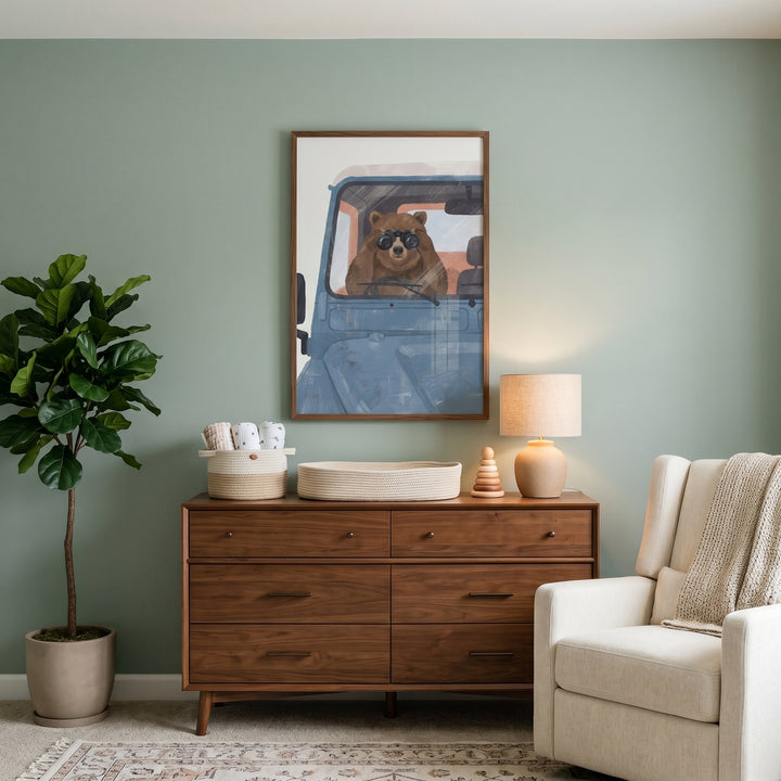Wood dresser below muted blue vehicle bear wall art on a green wall with a potted plant and a cream armchair.