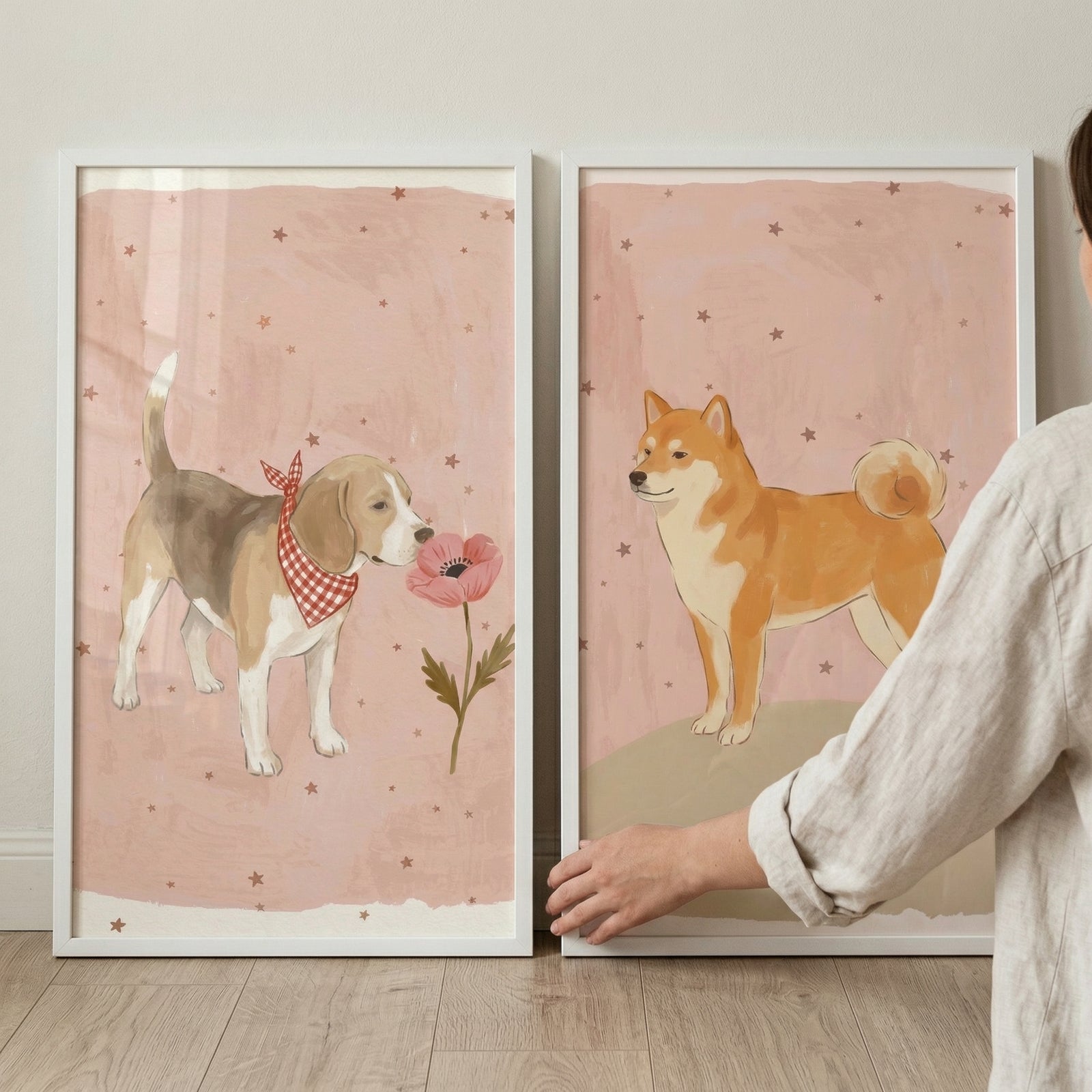 Two framed vertical illustrations sit on a light wood shelf against a neutral wall. Both pieces feature a soft, dusty pink background dotted with small brown stars. On the left, a tricolor hound-like dog wears a red-and-white checkered bandana and sniffs a single pink flower on a green stem. 