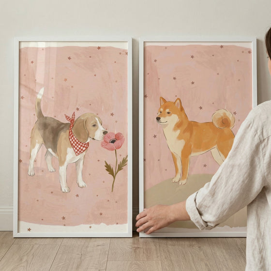 Two framed vertical illustrations sit on a light wood shelf against a neutral wall. Both pieces feature a soft, dusty pink background dotted with small brown stars. On the left, a tricolor hound-like dog wears a red-and-white checkered bandana and sniffs a single pink flower on a green stem. 
