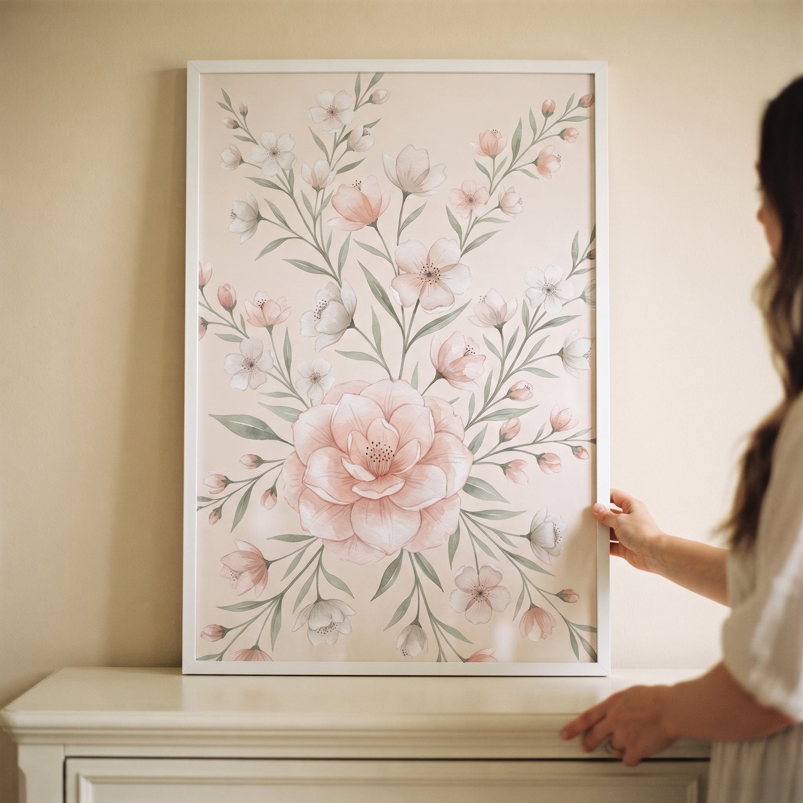 A person holds muted blush watercolor floral wall art in a white frame featuring delicate pink flowers and green leaves.