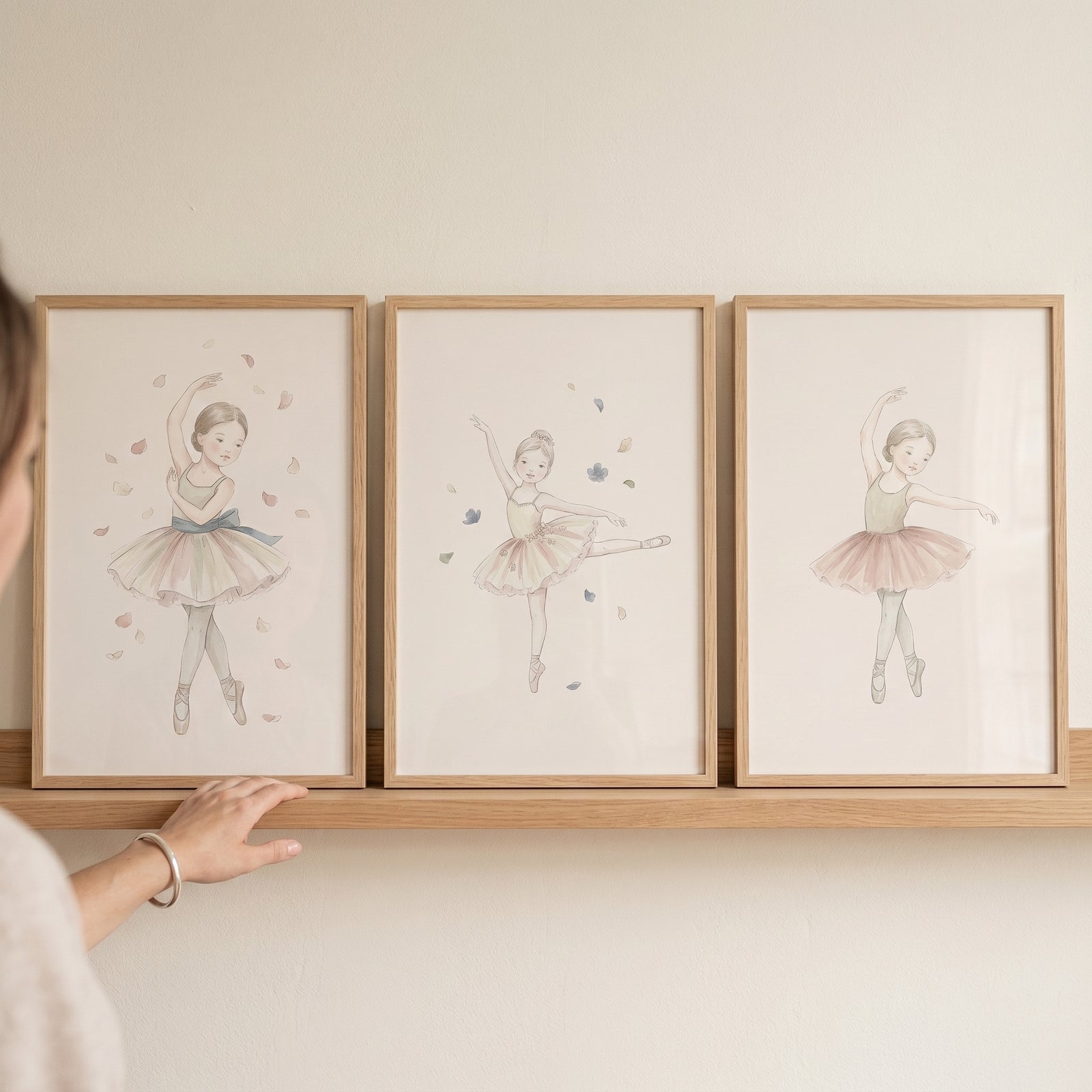A hand reaches toward a muted earthy tone ballerina wall art set of 3 in wooden frames on a shelf.