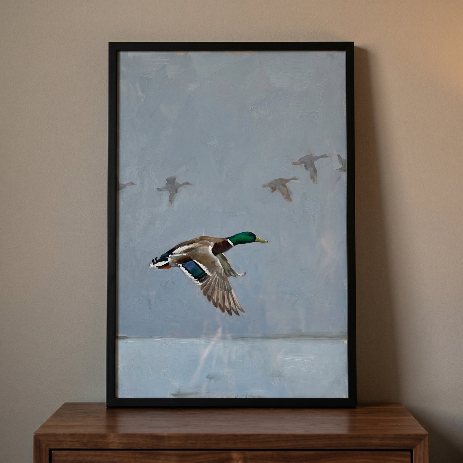 Muted flying mallard duck wall art in a black frame rests on a wooden dresser against a neutral wall.