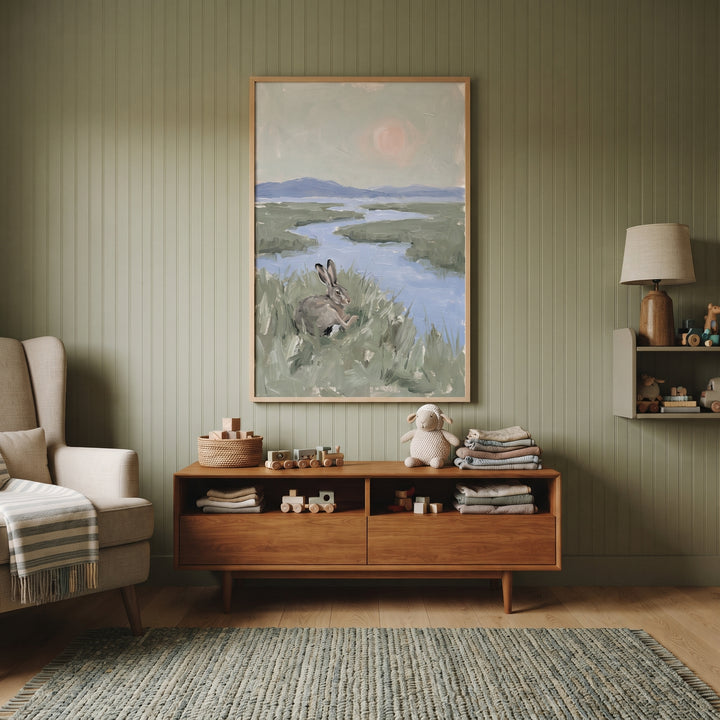 Muted marsh sitting bunny wall art hangs over a wooden console in a room with green paneled walls and a woven rug.