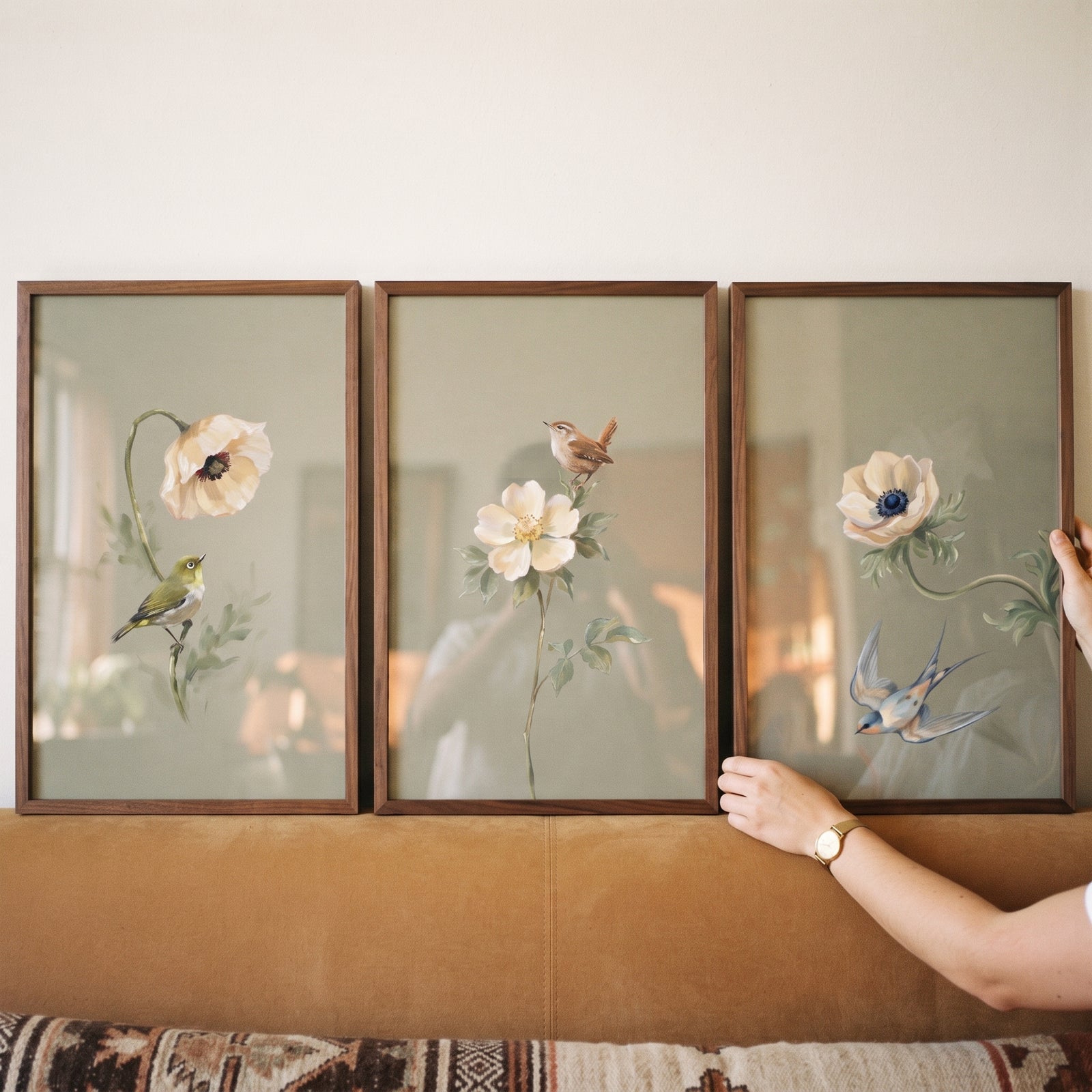 Muted neutral botanical bird print set of 3 in wood frames above a couch, with a hand adjusting one frame.
