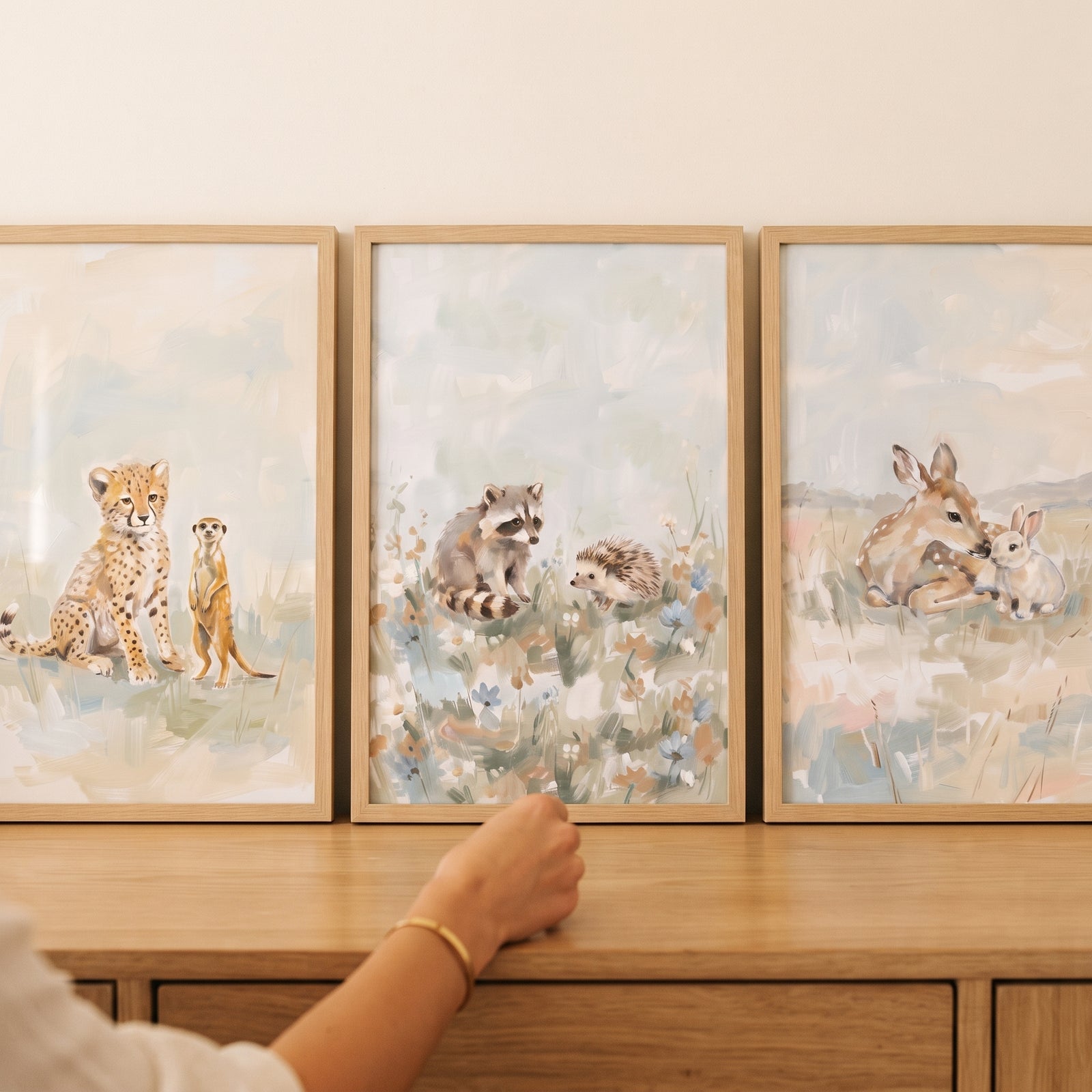 Hand touching a muted sage baby animals wall art set of 3 featuring a cheetah, raccoon, and deer on a wooden cabinet.