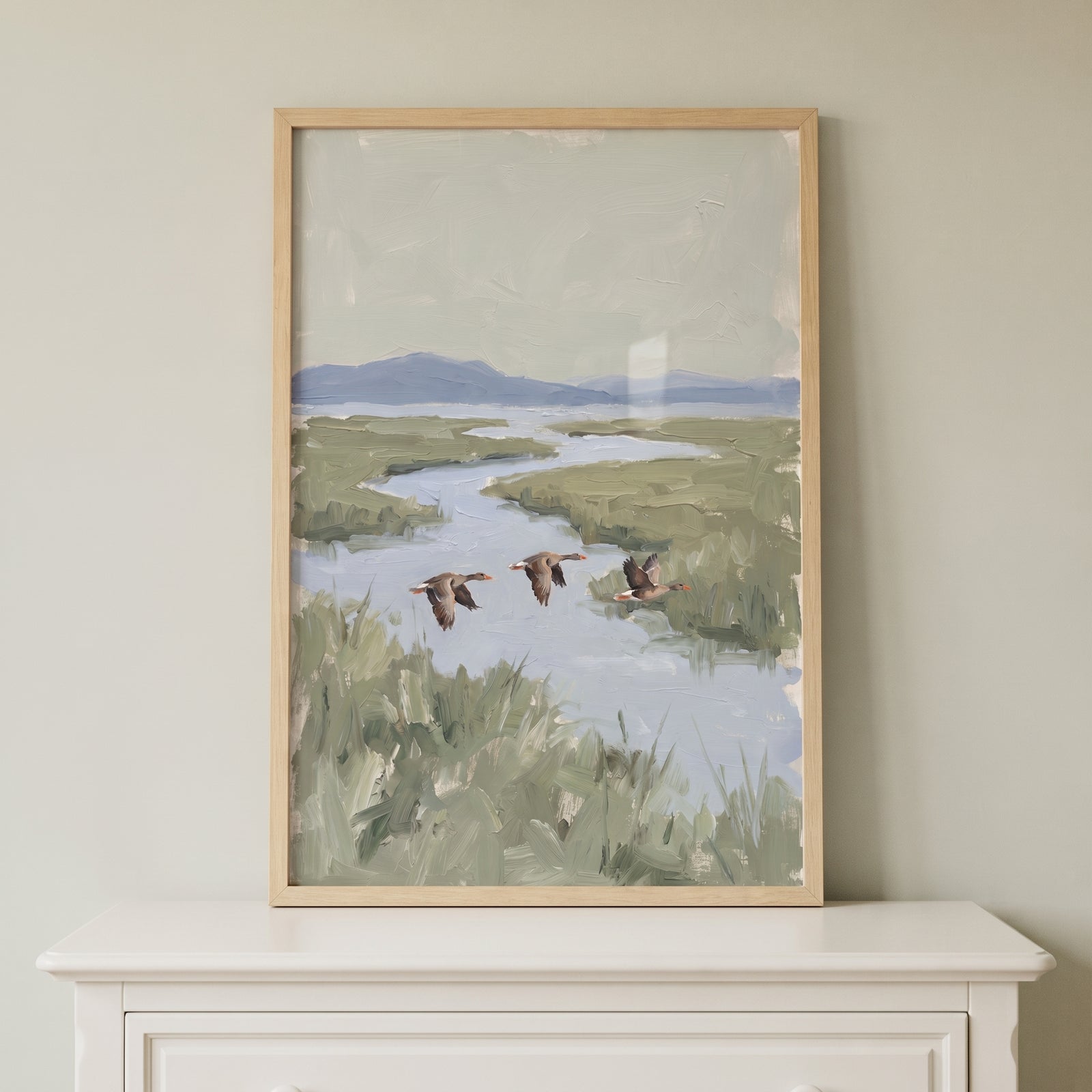 Muted sage flying geese artwork in a light frame hangs above a white dresser, showing birds over a winding river.