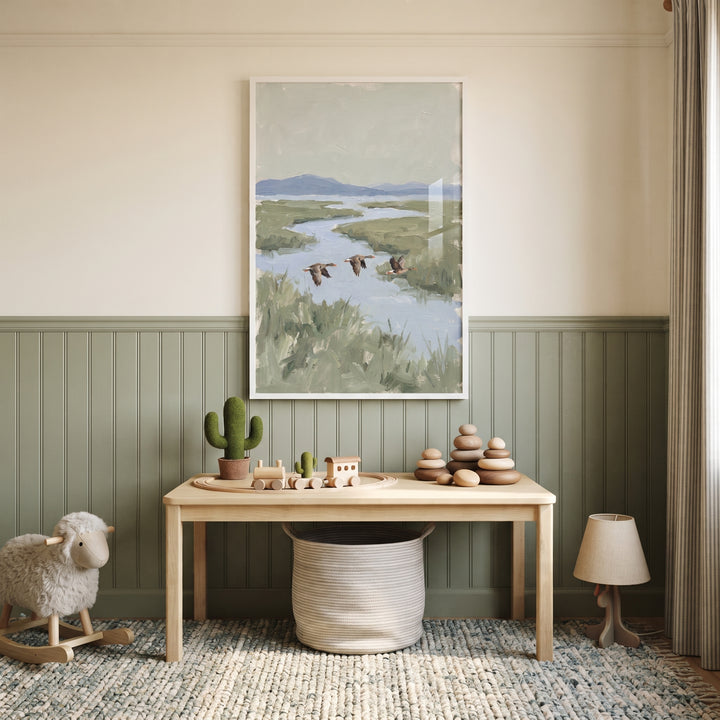 Muted sage flying geese artwork hangs above a wooden table. A rocking sheep and lamp sit on a blue rug in the green playroom.