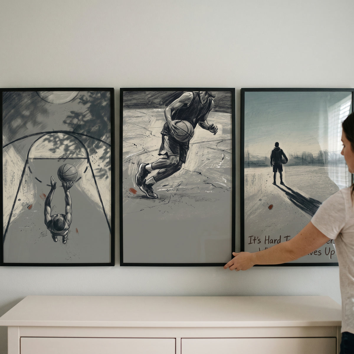 A muted sepia basketball player sports wall art set of 3 hangs on a wall above a dresser as someone adjusts the frames.