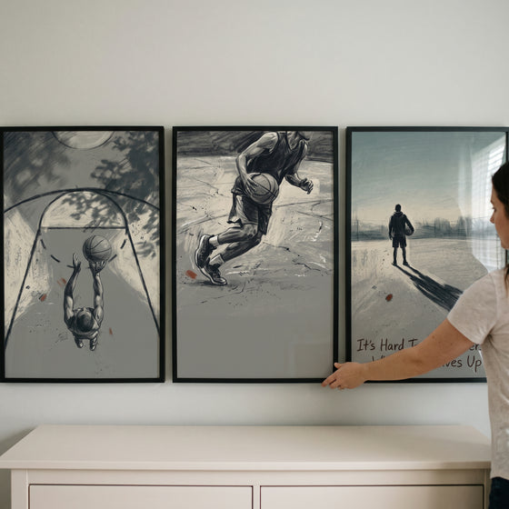 A muted sepia basketball player sports wall art set of 3 hangs on a wall above a dresser as someone adjusts the frames.