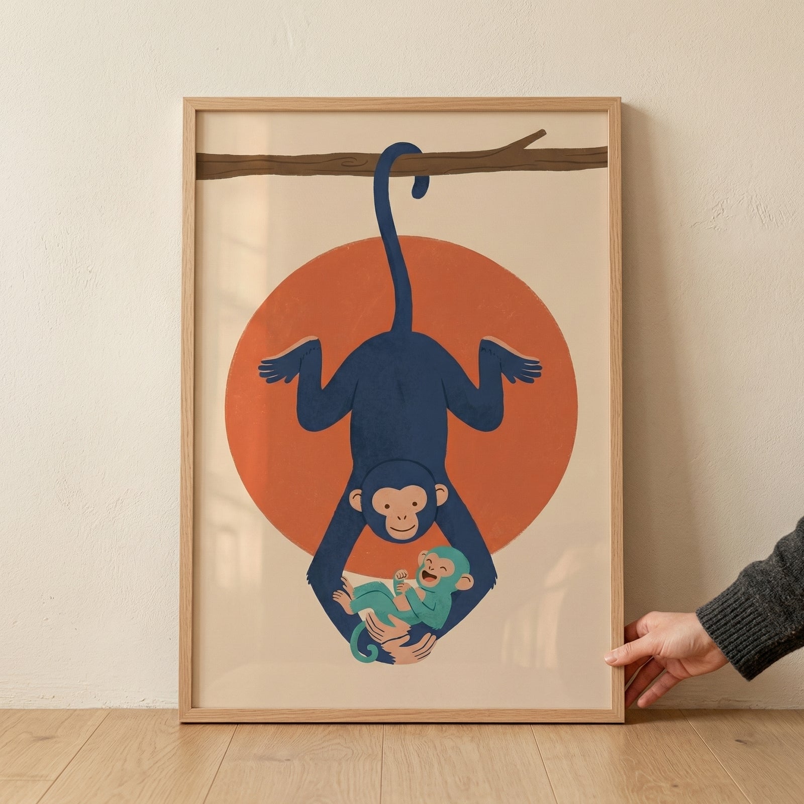 A minimalist graphic illustration, set within a light wood frame, depicts a large navy blue monkey hanging upside down from its tail on a simple brown branch. The monkey is smiling and cradling a small, teal-colored baby monkey in its long arms. Positioned behind the animals is a large, solid orange circle. The entire scene is set against a neutral cream background. The framed art is resting on a light wood floor against a white wall, with a hand holding its right edge.