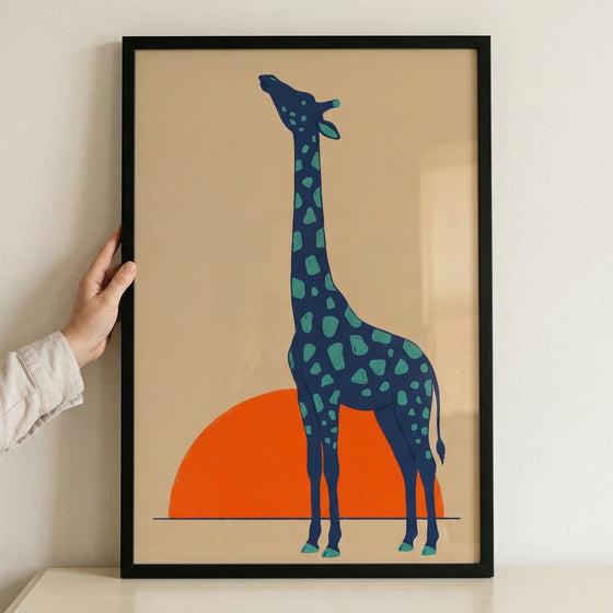 A framed graphic print of a stylized giraffe standing in front of a large orange sun. The giraffe is depicted in profile with a dark blue body and light green spots, looking upwards with its long neck. The entire scene is set against a solid tan background with a simple horizontal line representing the ground. The artwork is held by a hand on the left side and is enclosed in a minimalist black frame.