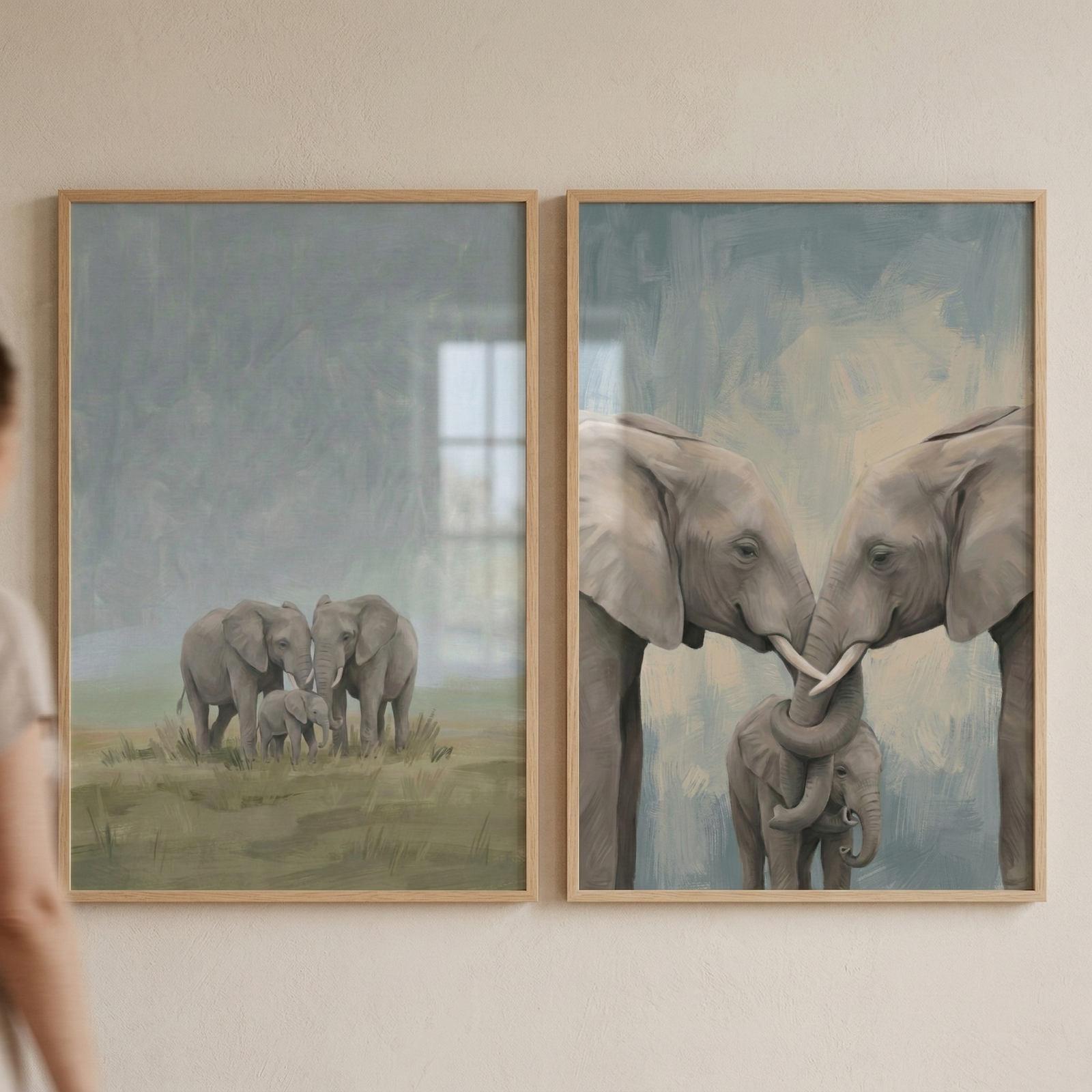 Two framed elephant paintings hang side by side on a light wall. One artwork shows a small elephant family standing together in soft grassy landscape with a baby elephant between two adults. The second shows two adult elephants gently touching trunks above a baby elephant standing between them.