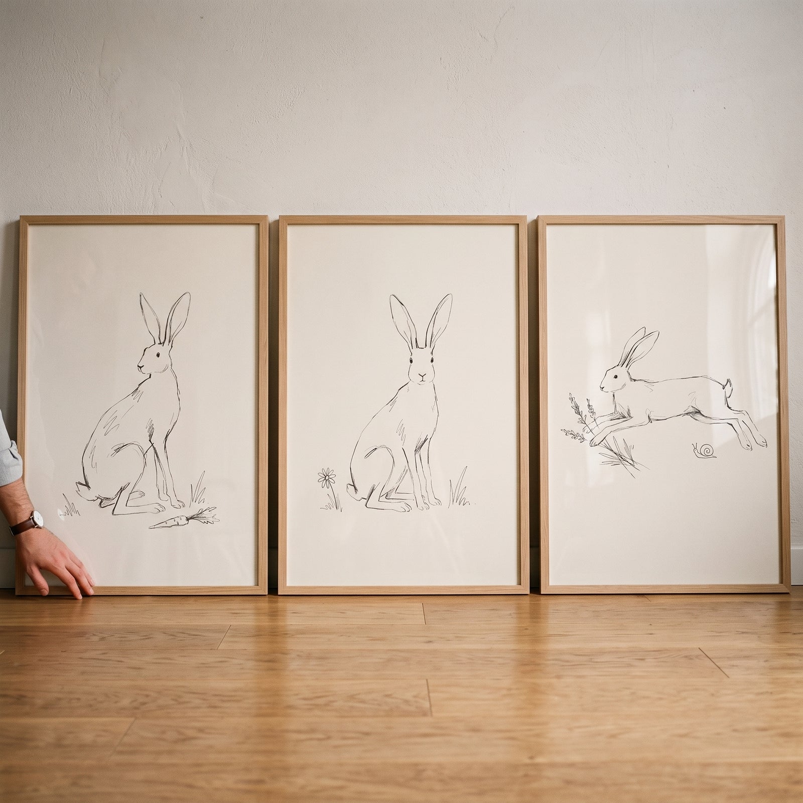 Neutral bunny wall art set of 3 in light wood frames leaning against a white wall on a wooden floor.