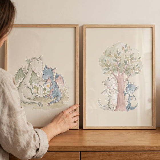 Two watercolor dragons read a book and hide behind a tree in this neutral pastel baby dragons wall art set of 2.