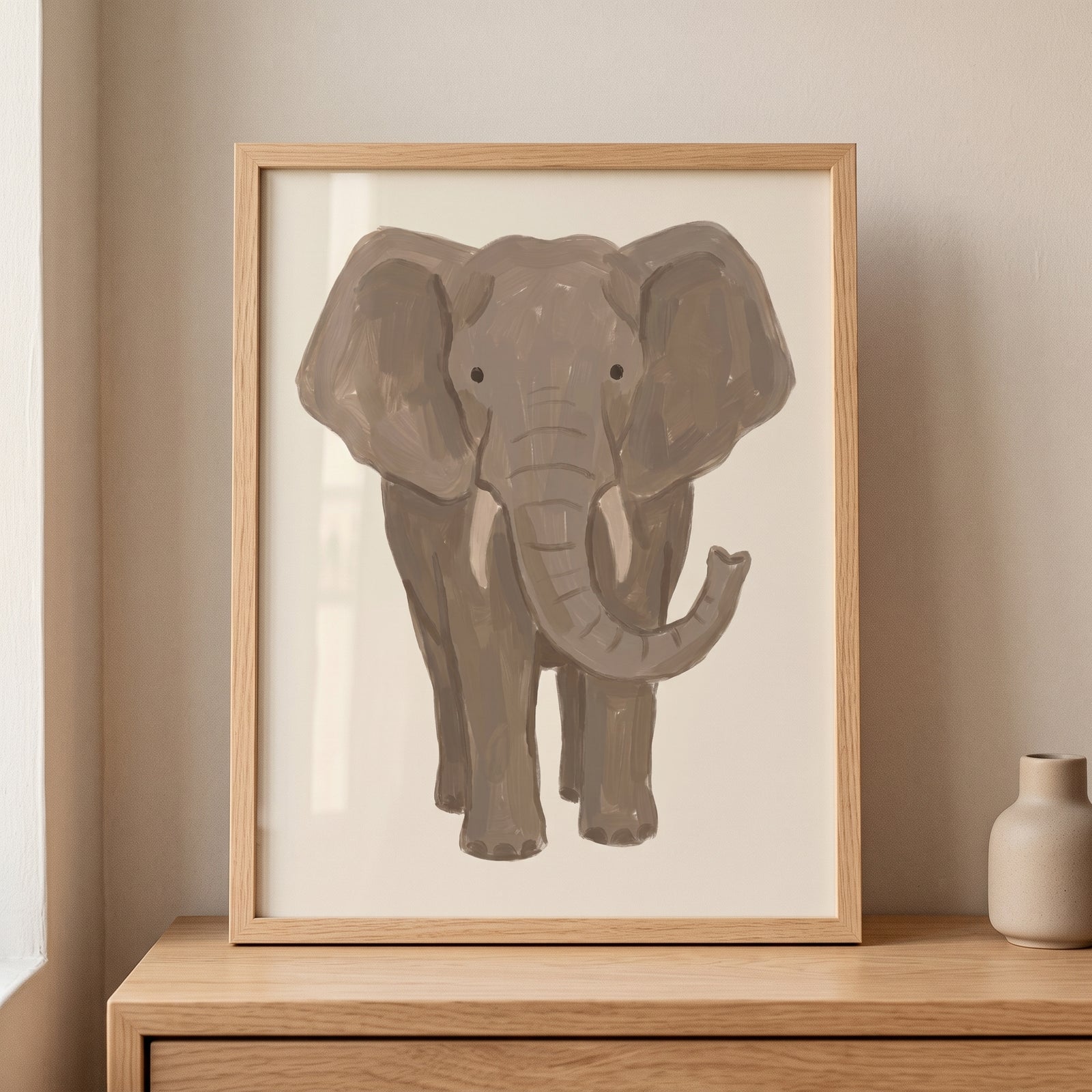 Framed neutral tan baby elephant wall art sits on a wooden dresser next to a small beige vase.