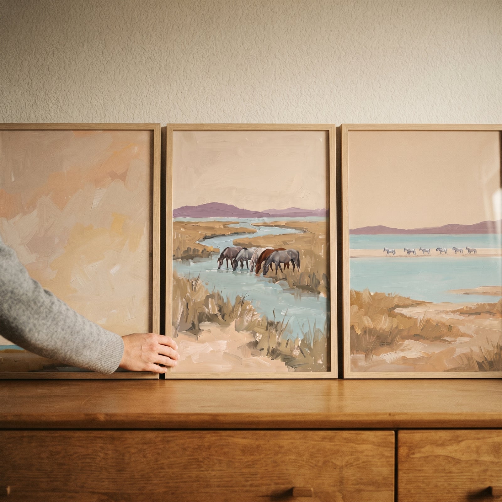 Hand adjusting a neutral tan coastal horses wall art set of 3 displayed on a wooden dresser against a cream wall.