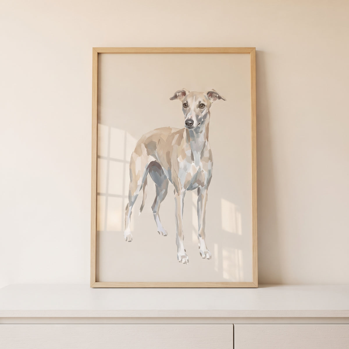 Neutral tan italian greyhound wall art in a light wood frame sits on a white surface against a beige wall.