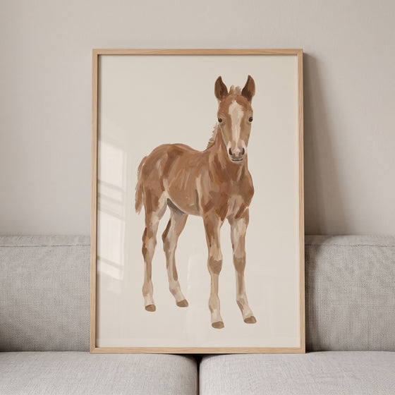 Neutral tan standing horse wall art featuring a brown foal with white markings in a wood frame on a gray sofa.