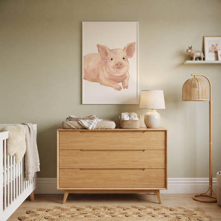Neutral tone smiling pig wall art hangs above a wooden dresser in a soft green nursery with a woven floor lamp.